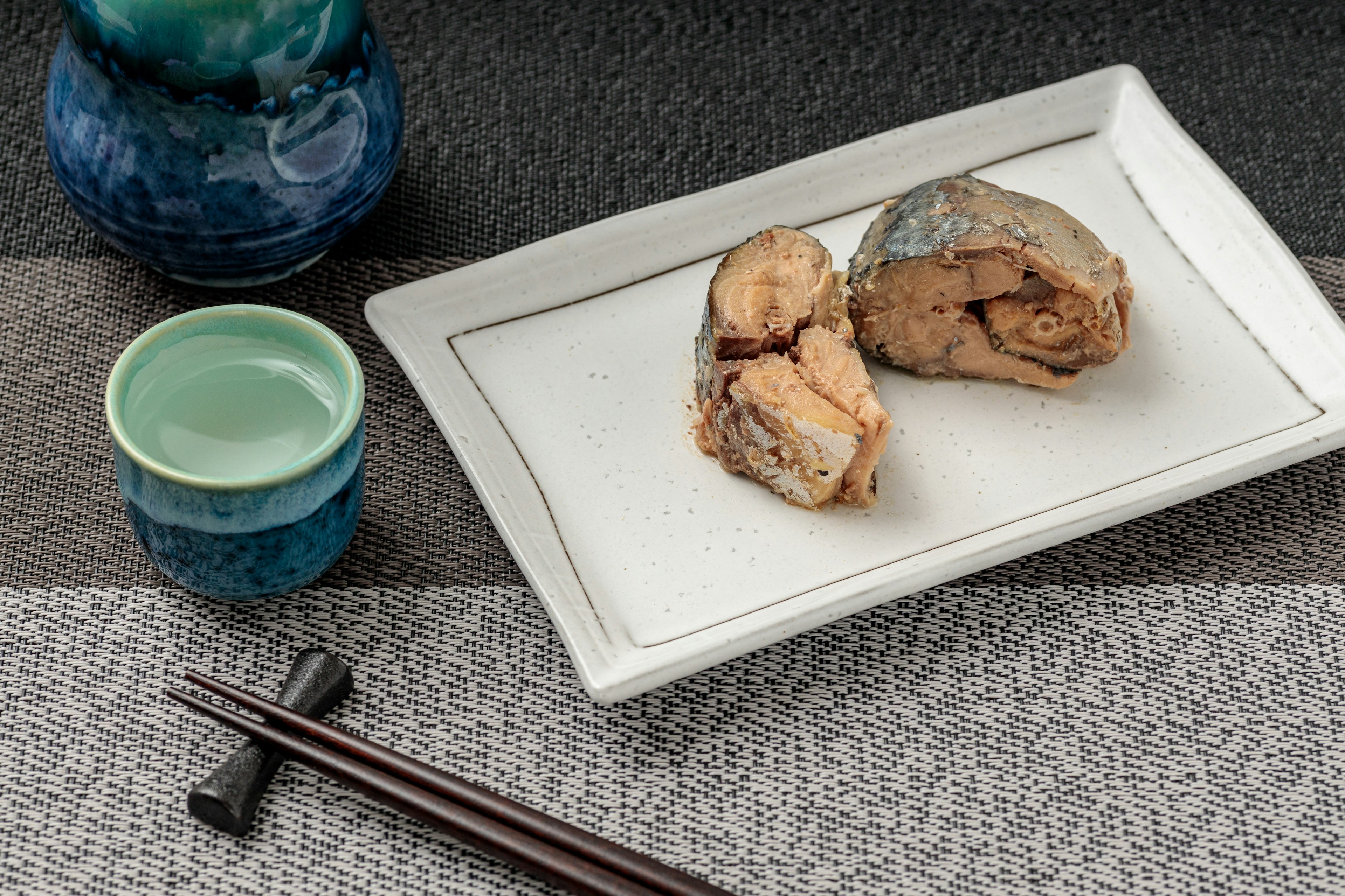 Poisson grillé servi sur une assiette blanche avec une tasse à thé verte et une bouteille de saké bleue