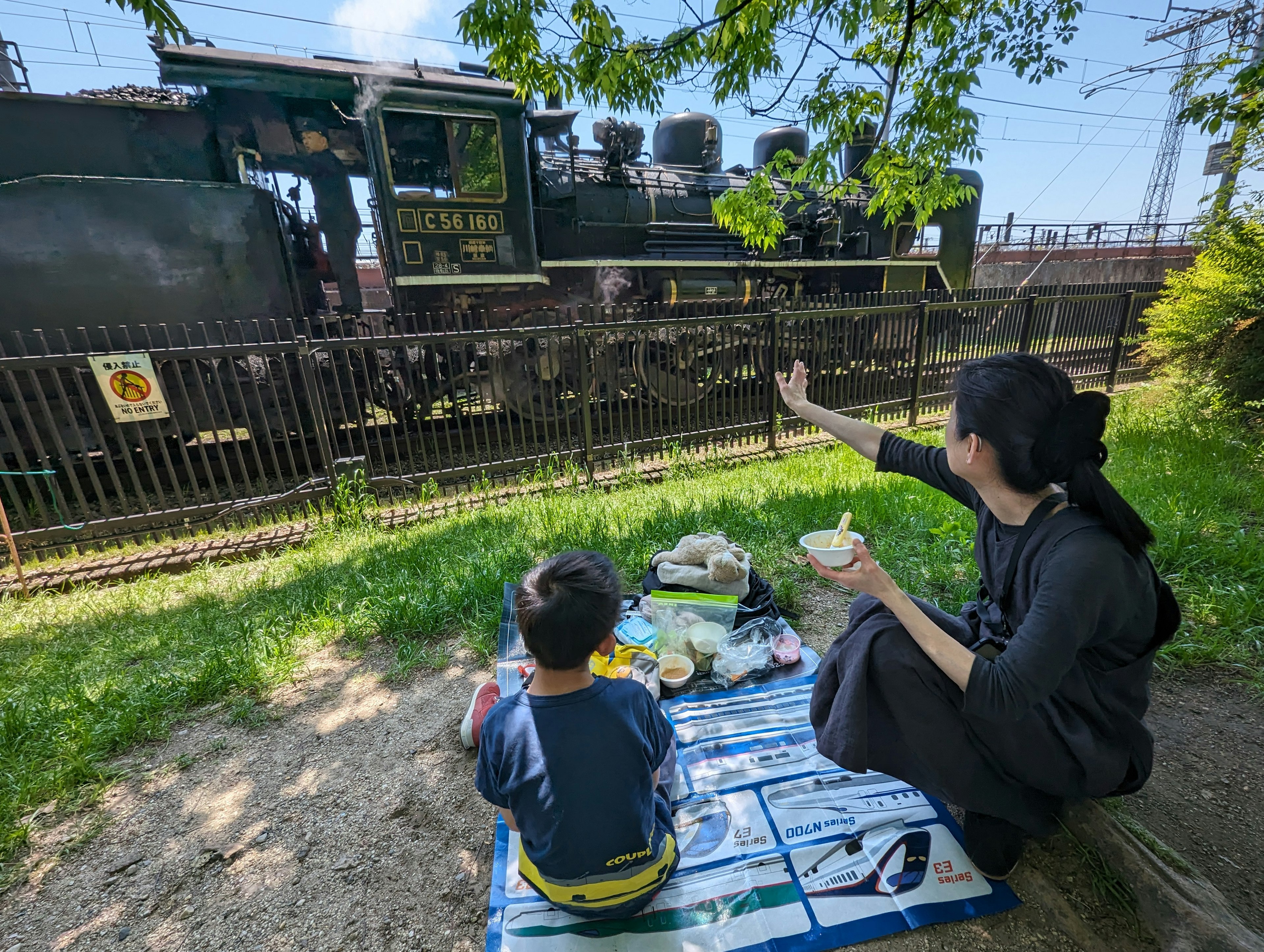 Một người mẹ và đứa trẻ đang thưởng thức bữa ăn dã ngoại trong khi một chiếc đầu máy hơi nước đi qua