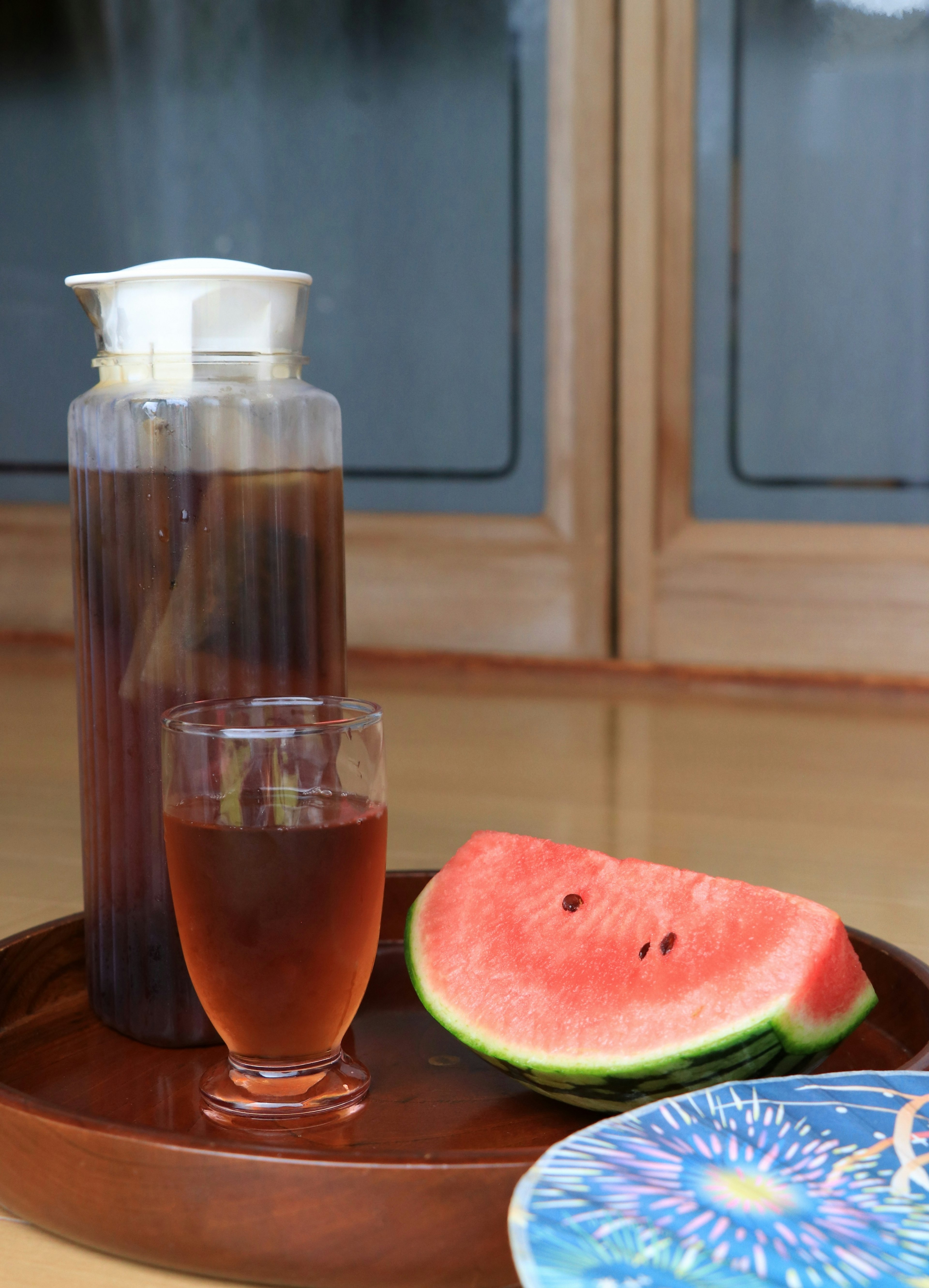 A glass pitcher with a drink and cup alongside a slice of watermelon on a blue plate