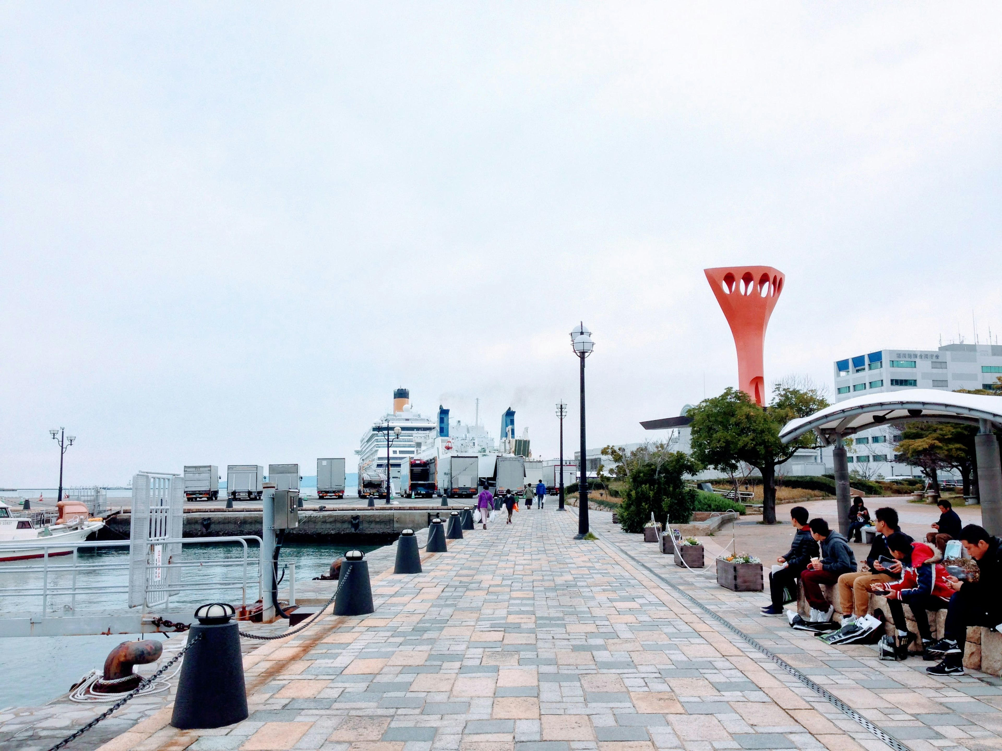 海邊步道上坐著的人和港口景觀，紅色塔樓和船隻