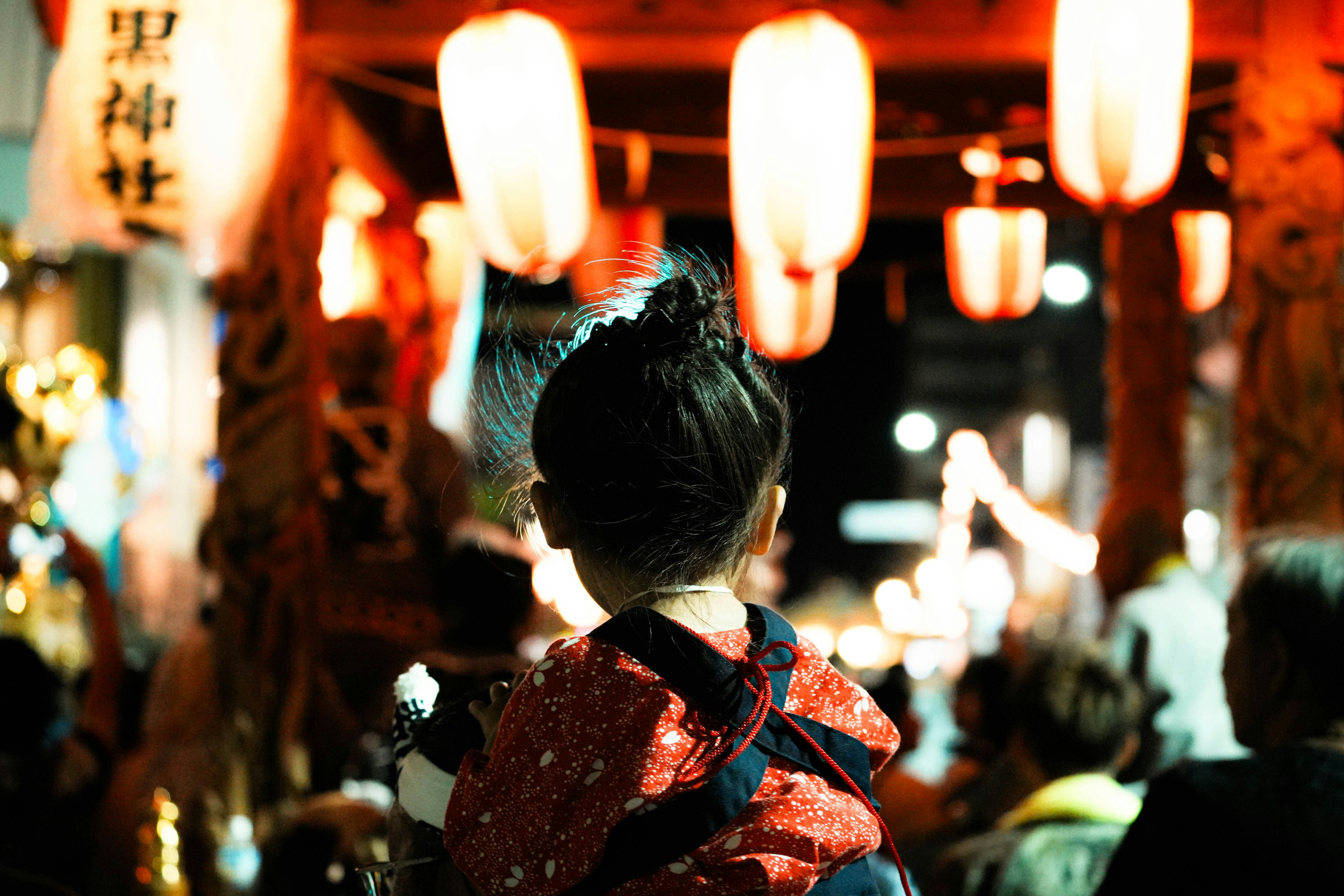 Seorang anak berpakaian kimono merah menikmati festival dengan lampion di latar belakang