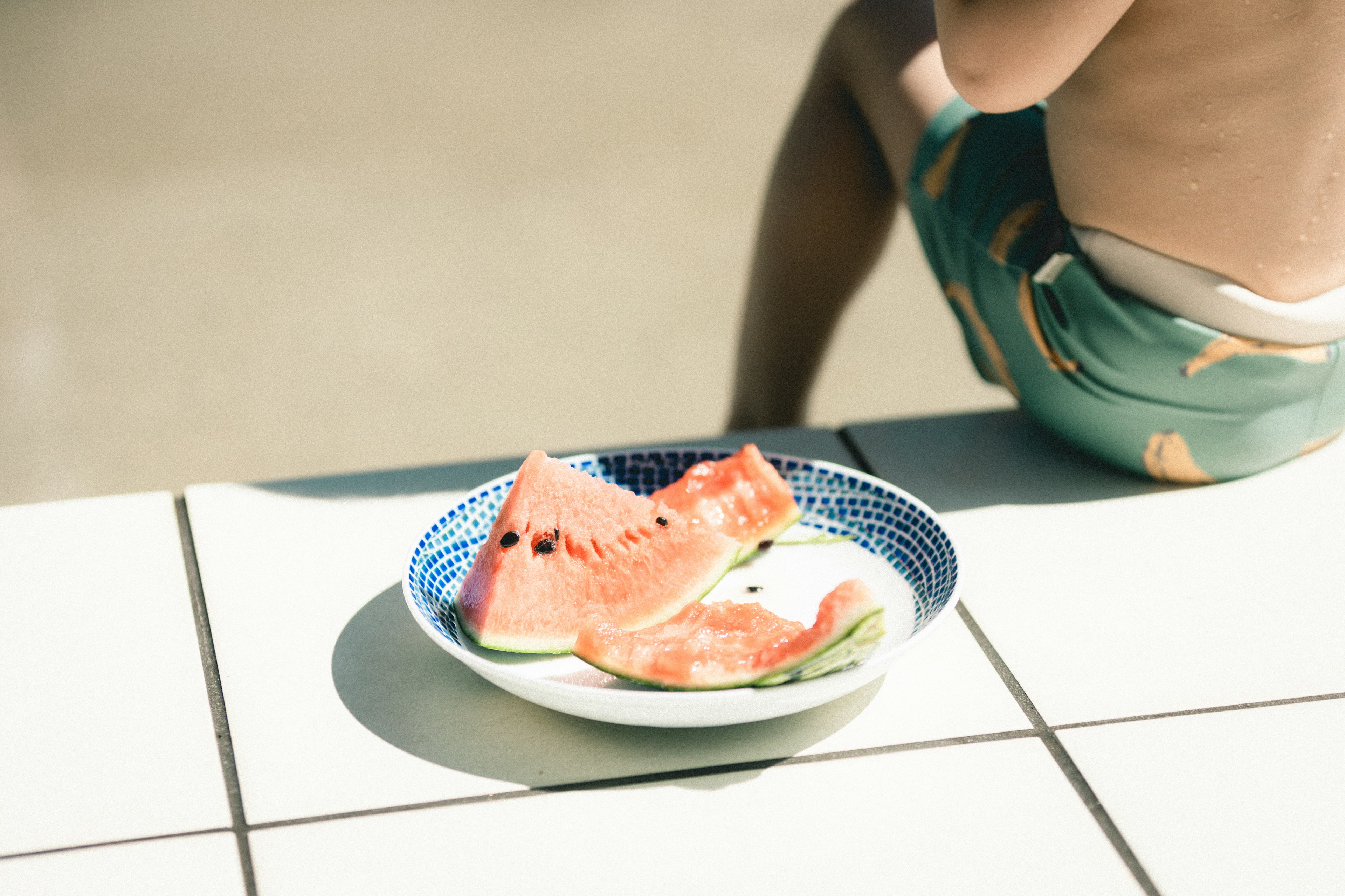 Un plato con rodajas de sandía al borde de la piscina con un niño sentado cerca