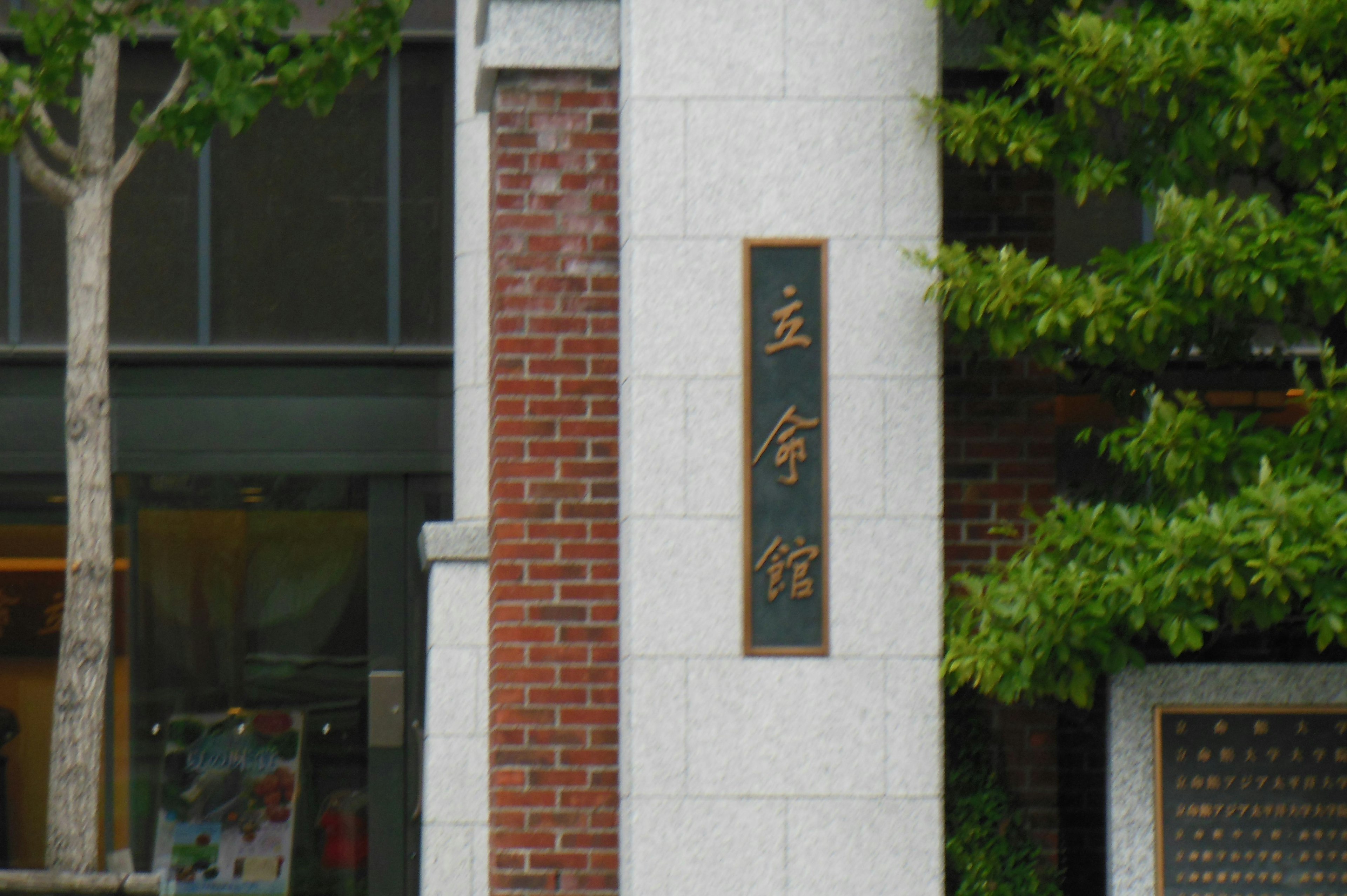 Exterior de un edificio con una columna de piedra y árboles verdes, letrero negro con caracteres blancos que dice '主命館'