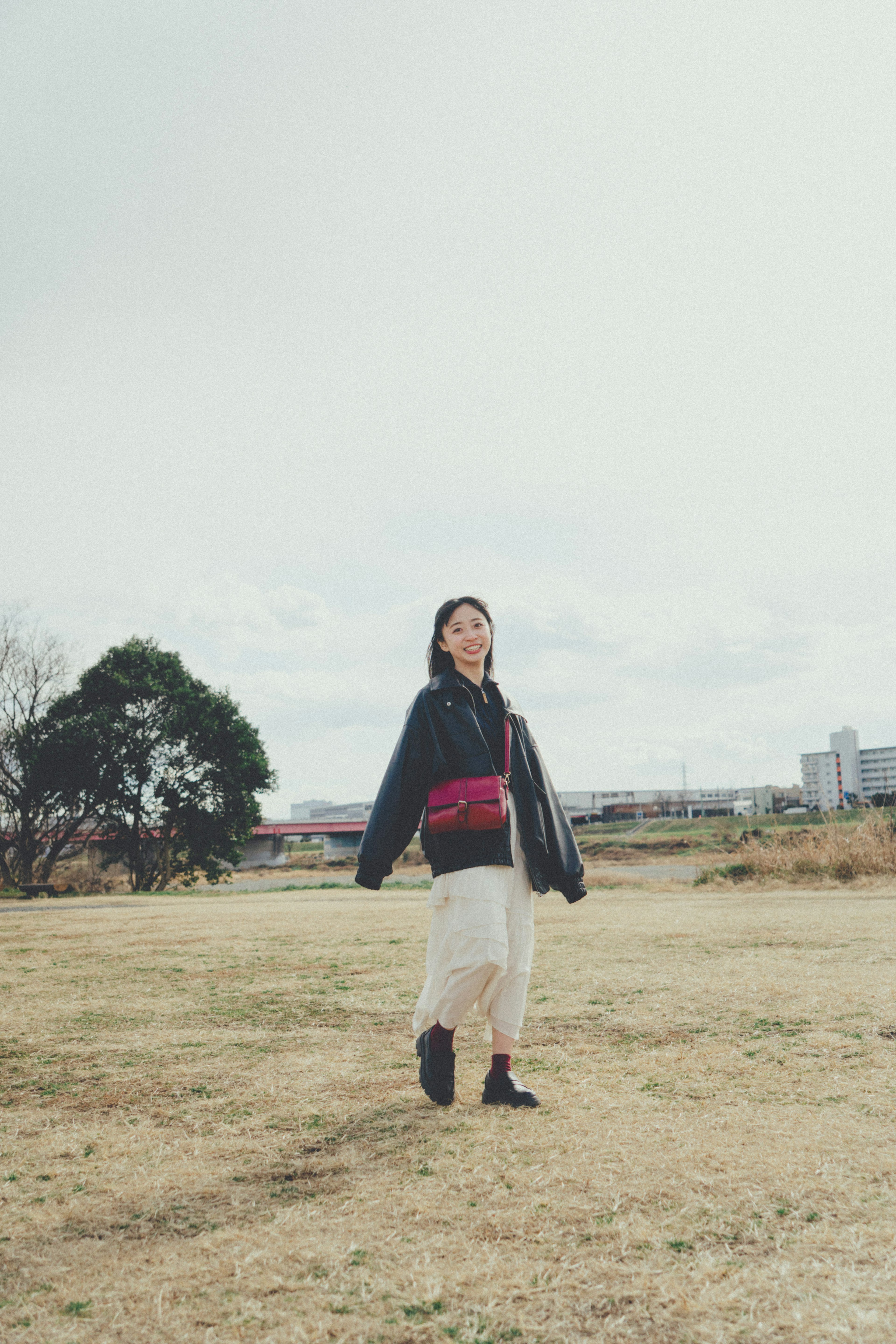 A woman standing in a vast grassy field wearing a black jacket and white skirt holding a red bag