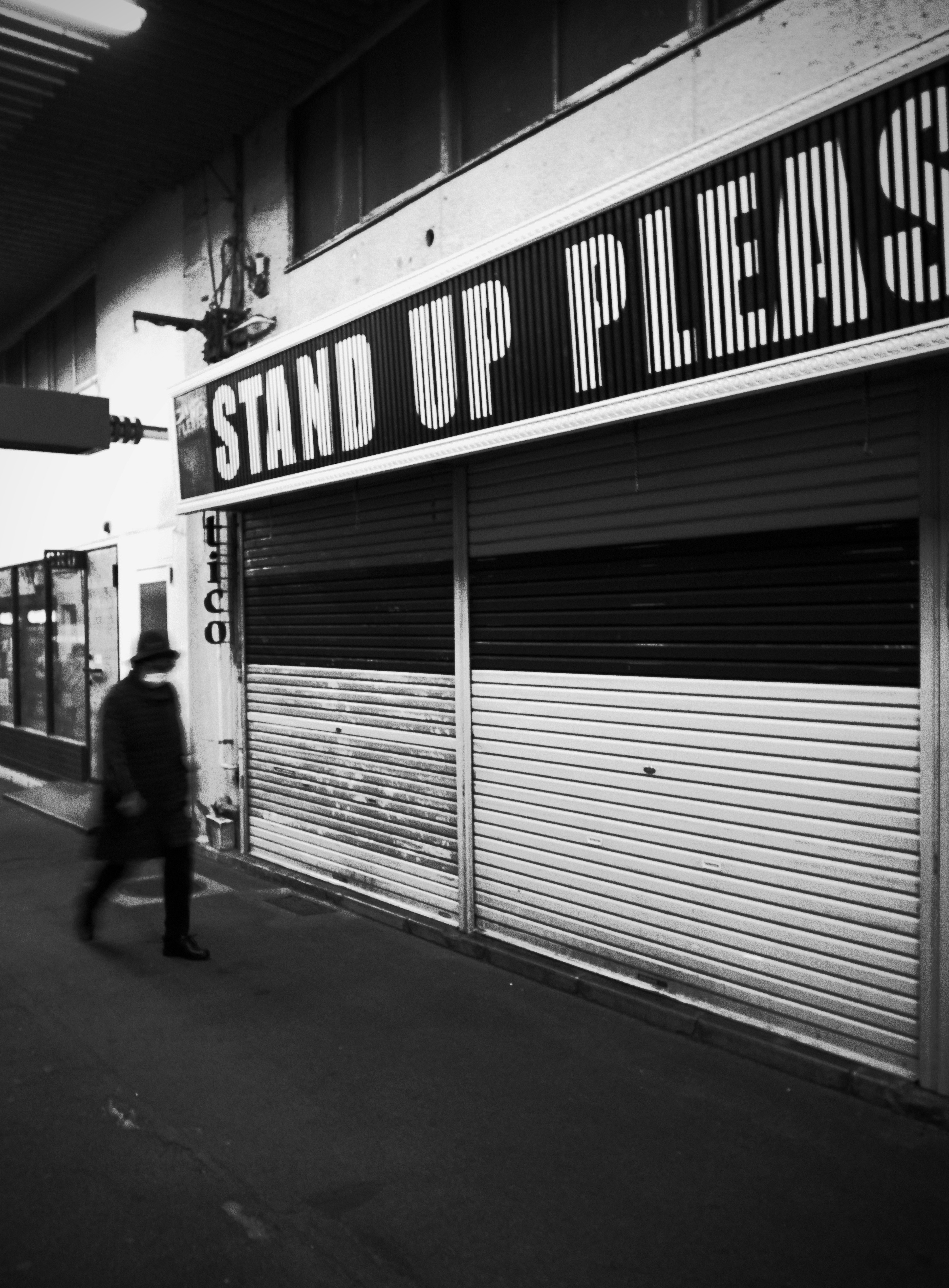 黒白の街中で歩く人物と「STAND UP PLEASE」の看板