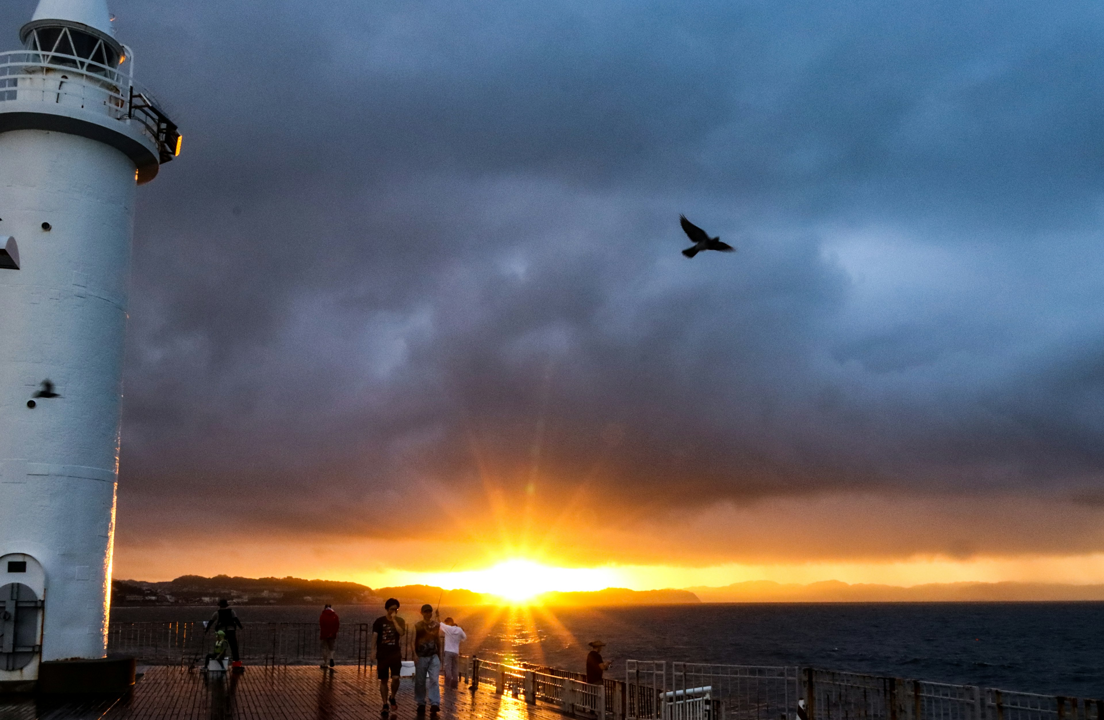 A lighthouse with people silhouetted against a sunset over the ocean