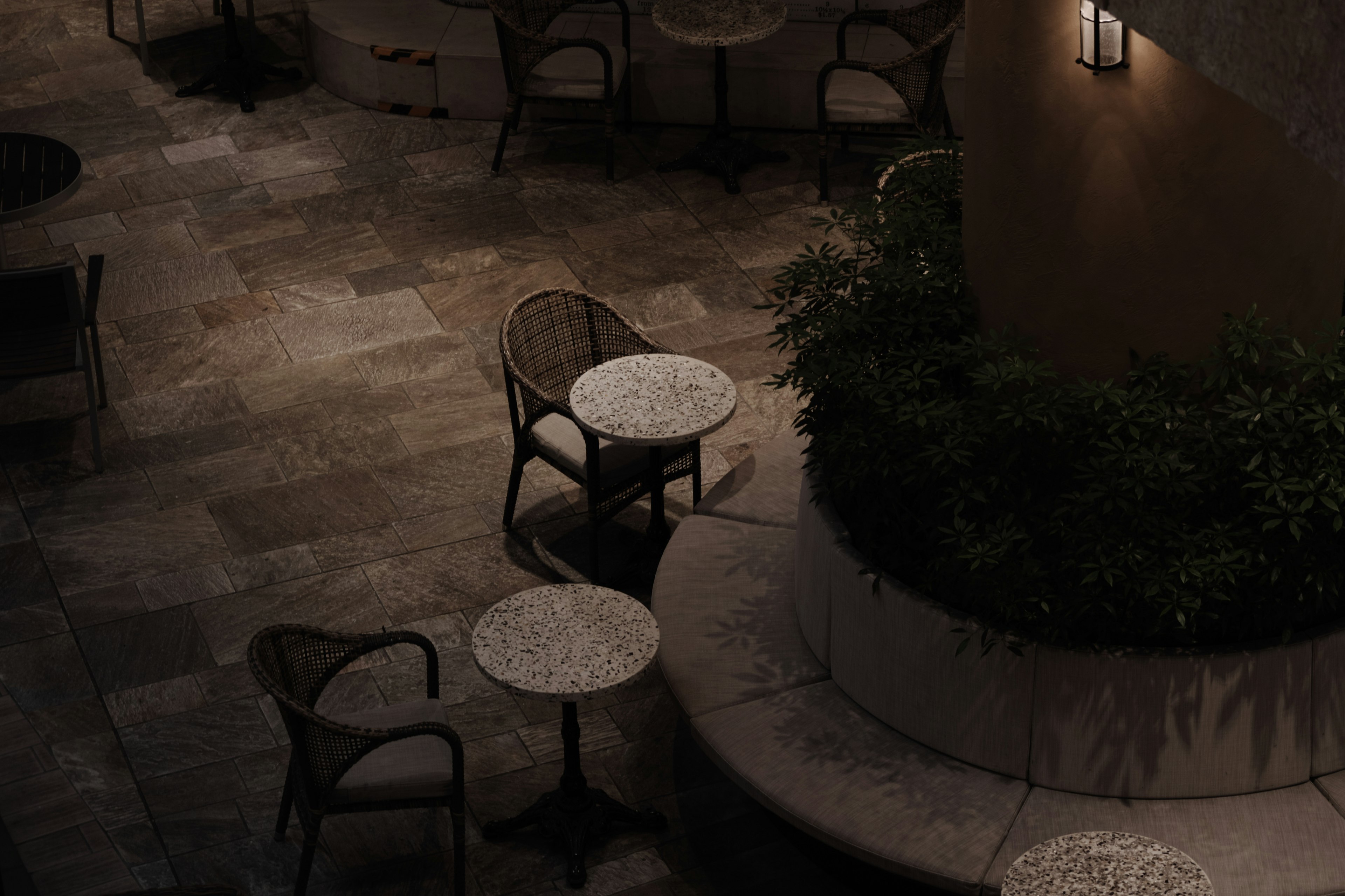 A dimly lit outdoor seating area with round tables and chairs surrounded by greenery