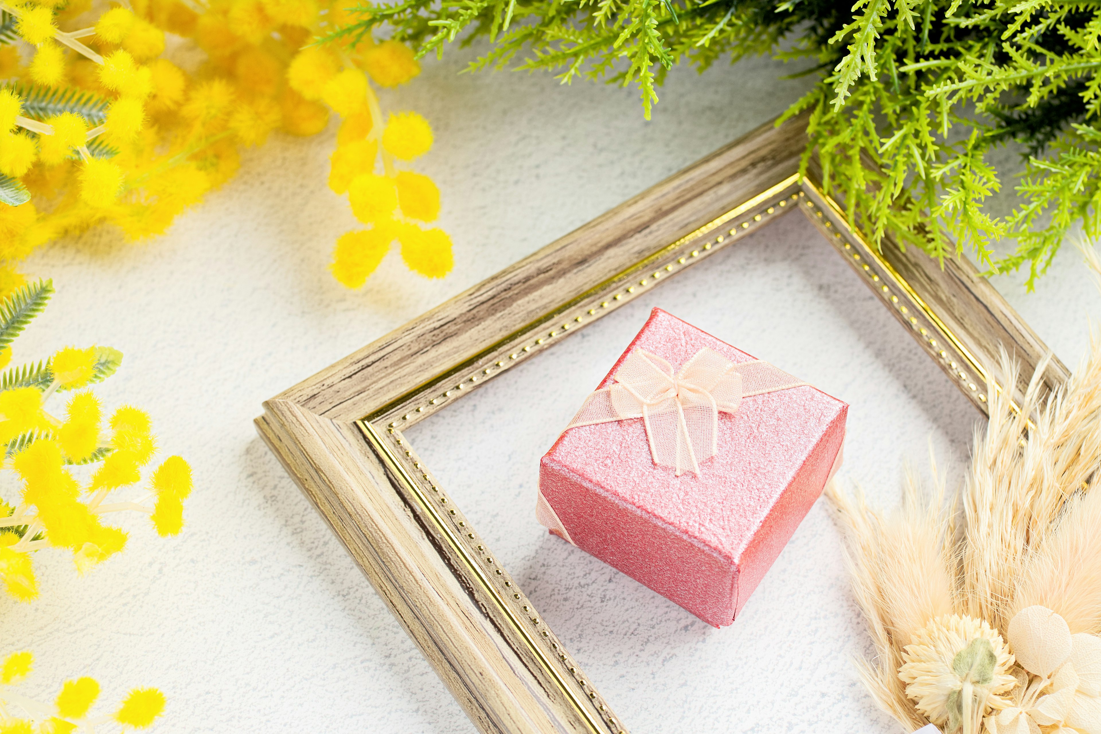 A pink gift box surrounded by yellow flowers and dried plants within a decorative frame