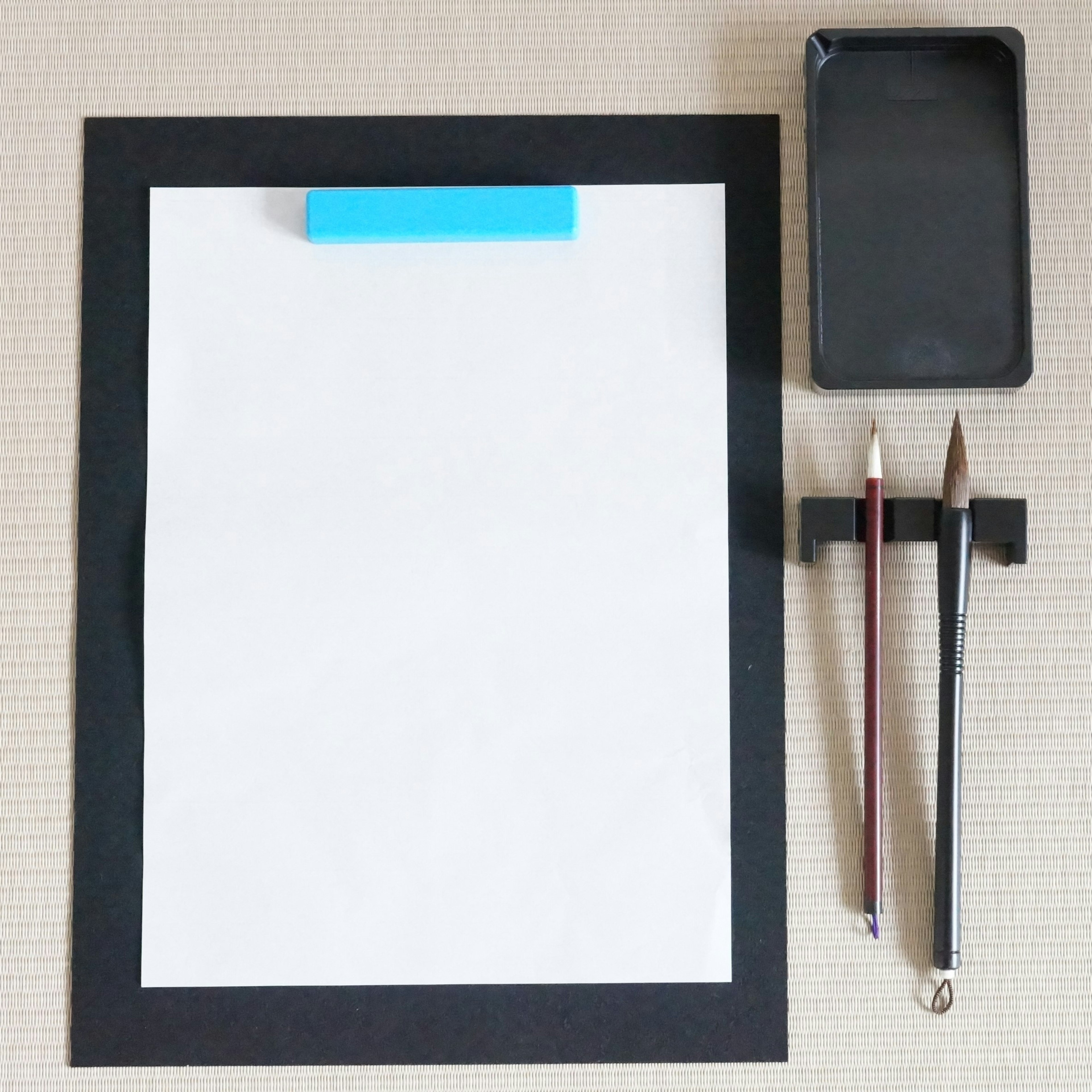 A blank sheet of paper with a blue clip on a black frame accompanied by a black smartphone and two pencils