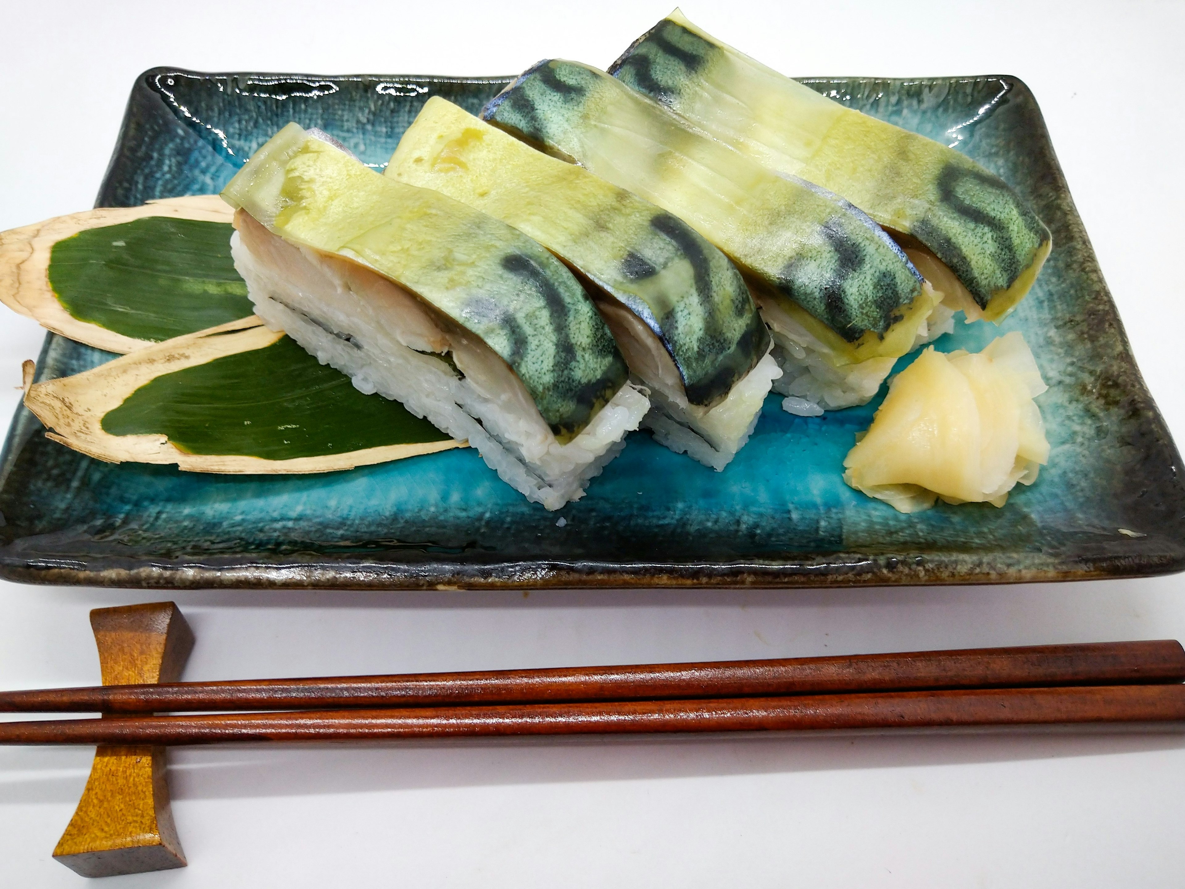 Pressed sushi with mackerel served on a blue plate with ginger and shiso leaves