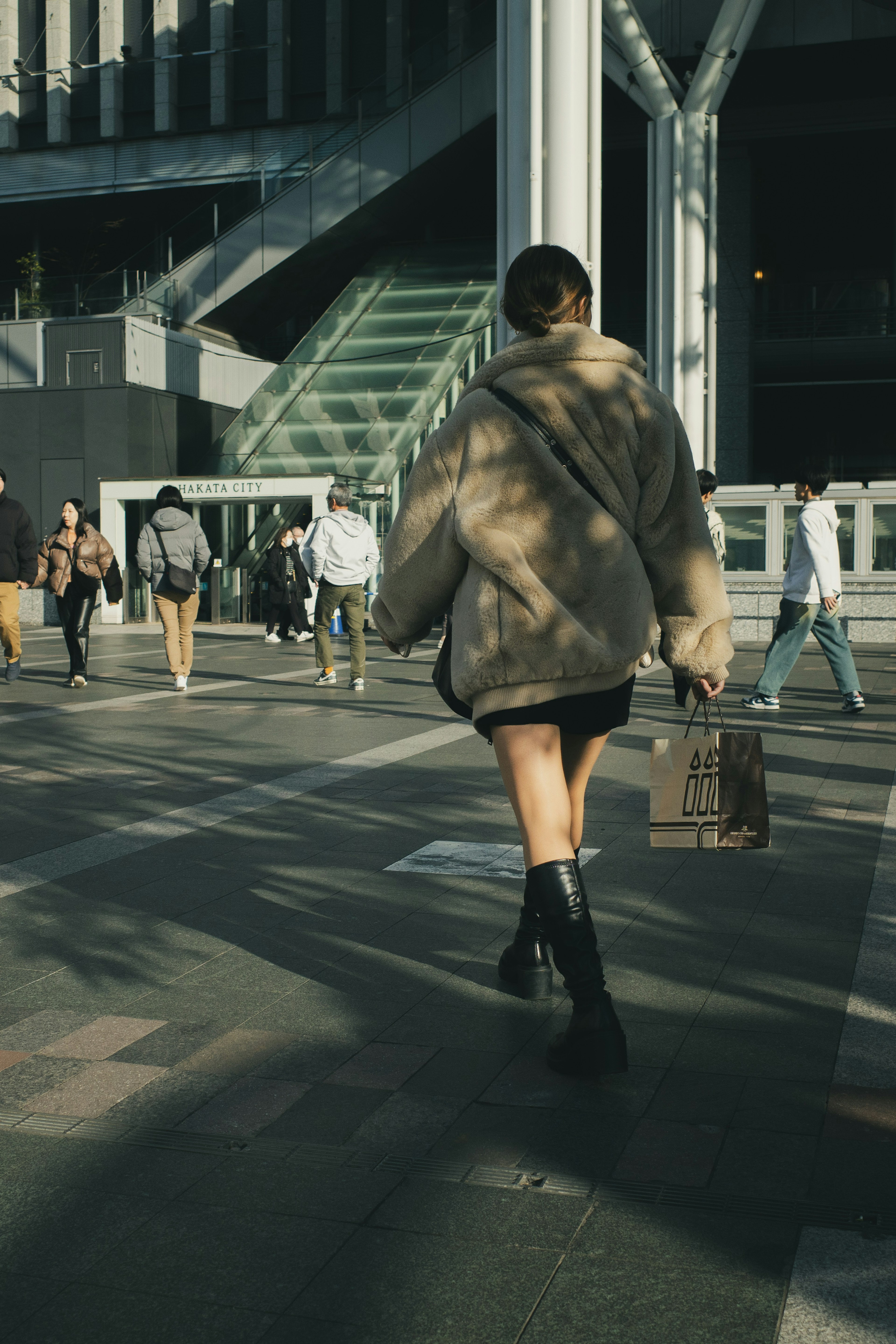 Woman walking outdoors seen from behind wearing a large coat and black boots surrounded by other people