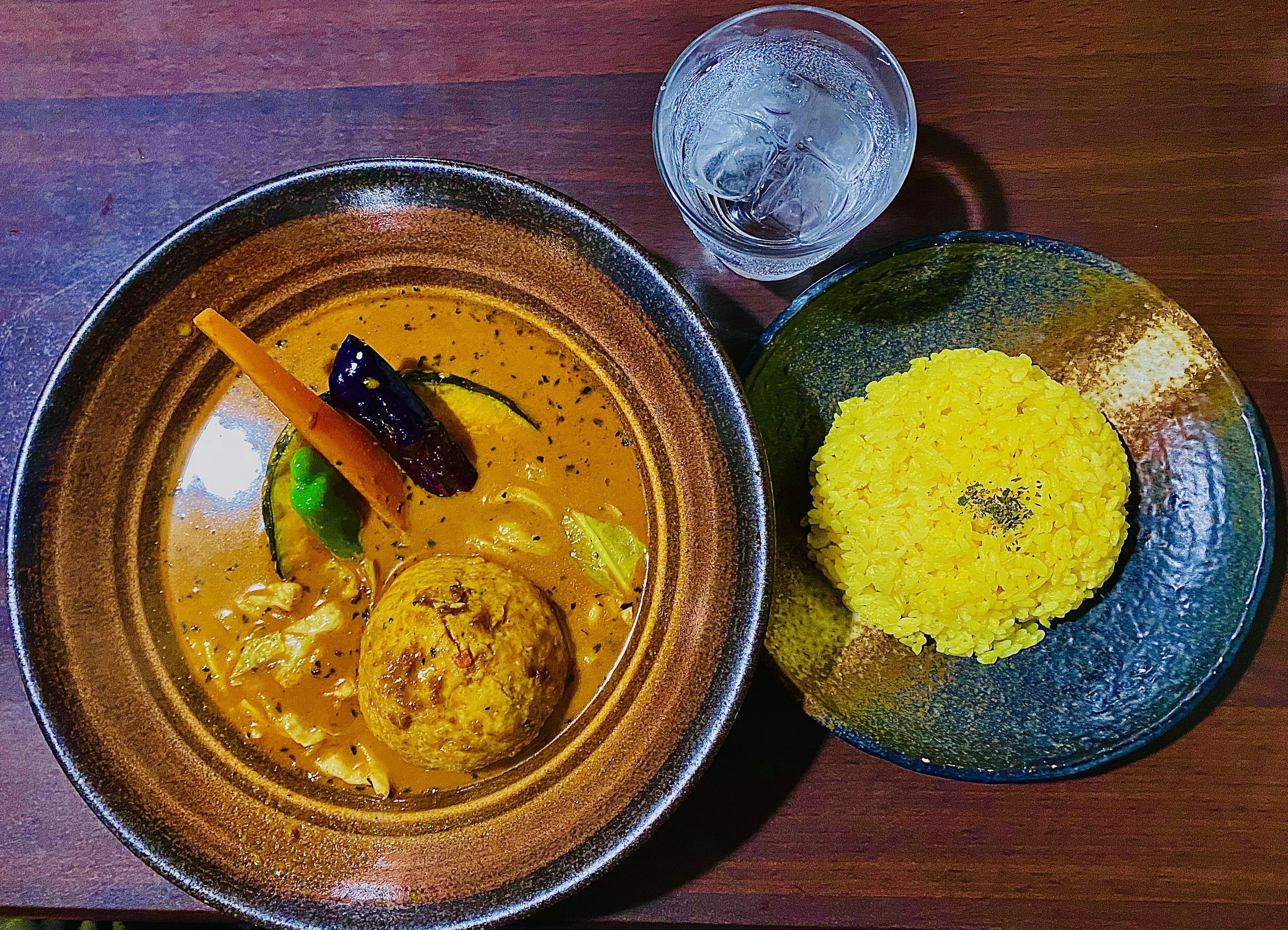 A bowl of curry with vegetables and two rice balls served with a glass of water