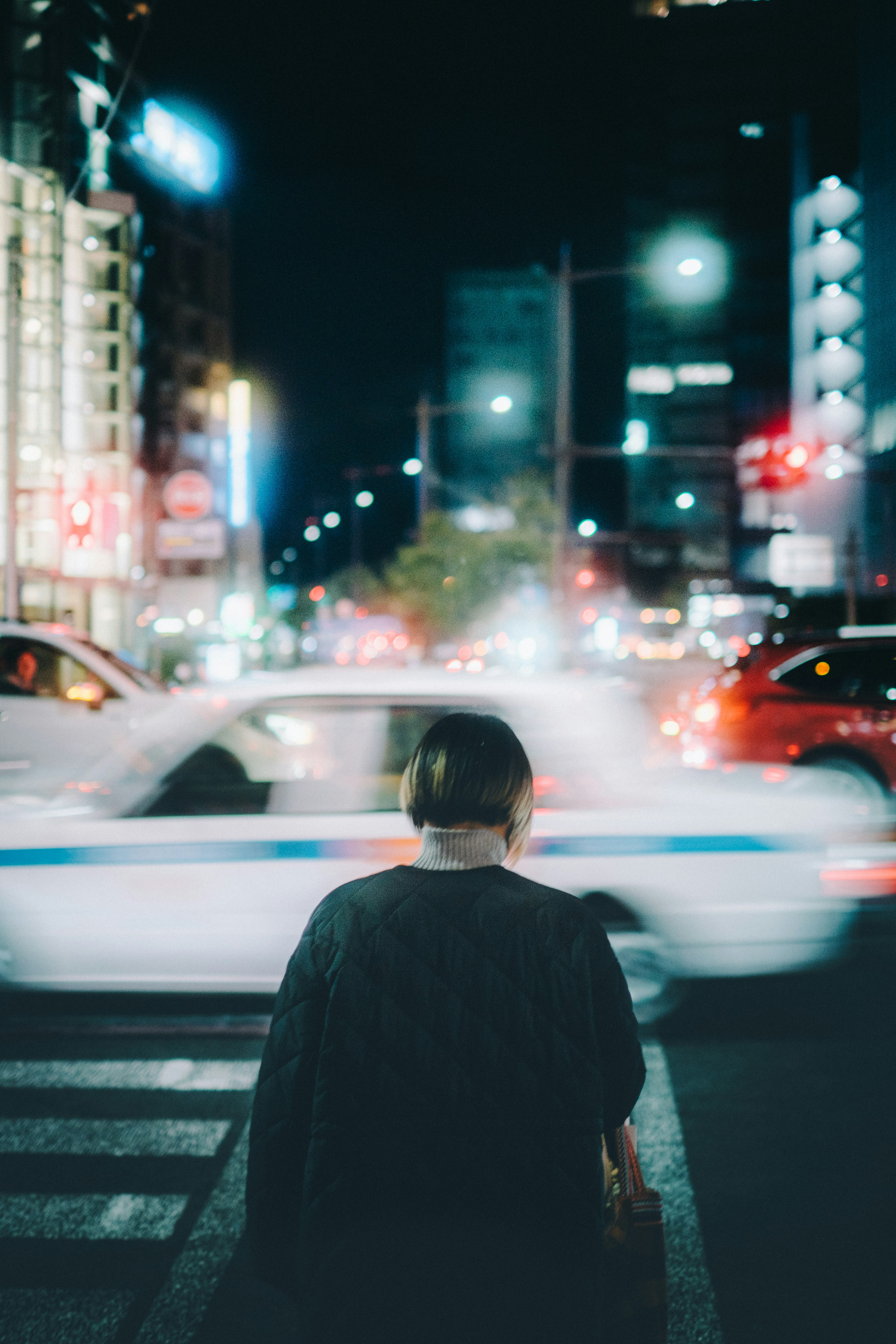 Person standing at a crosswalk with a taxi passing by in a city at night