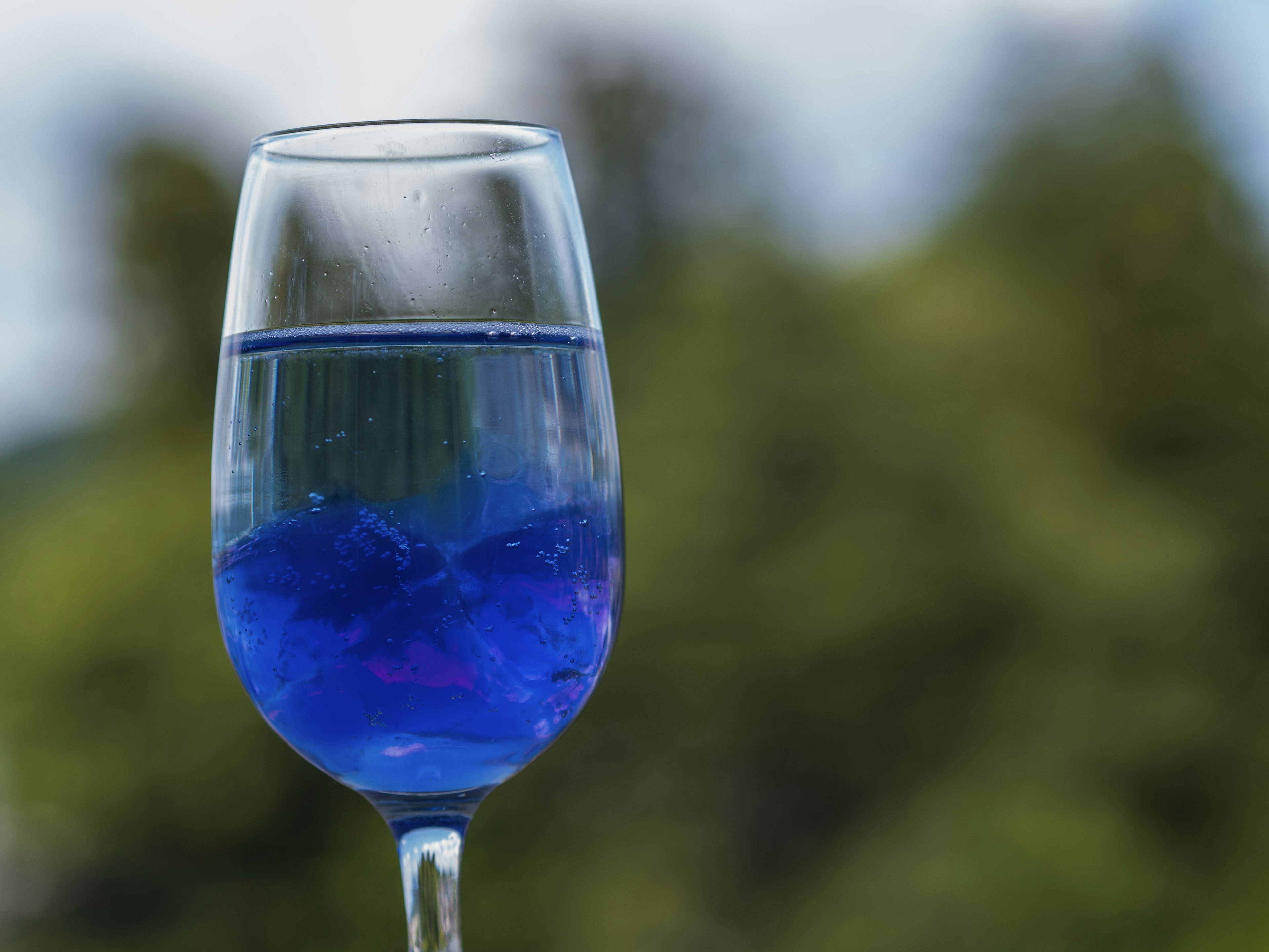 Ein transparentes Glas mit blauer Flüssigkeit und Eiswürfeln vor einem verschwommenen grünen Hintergrund