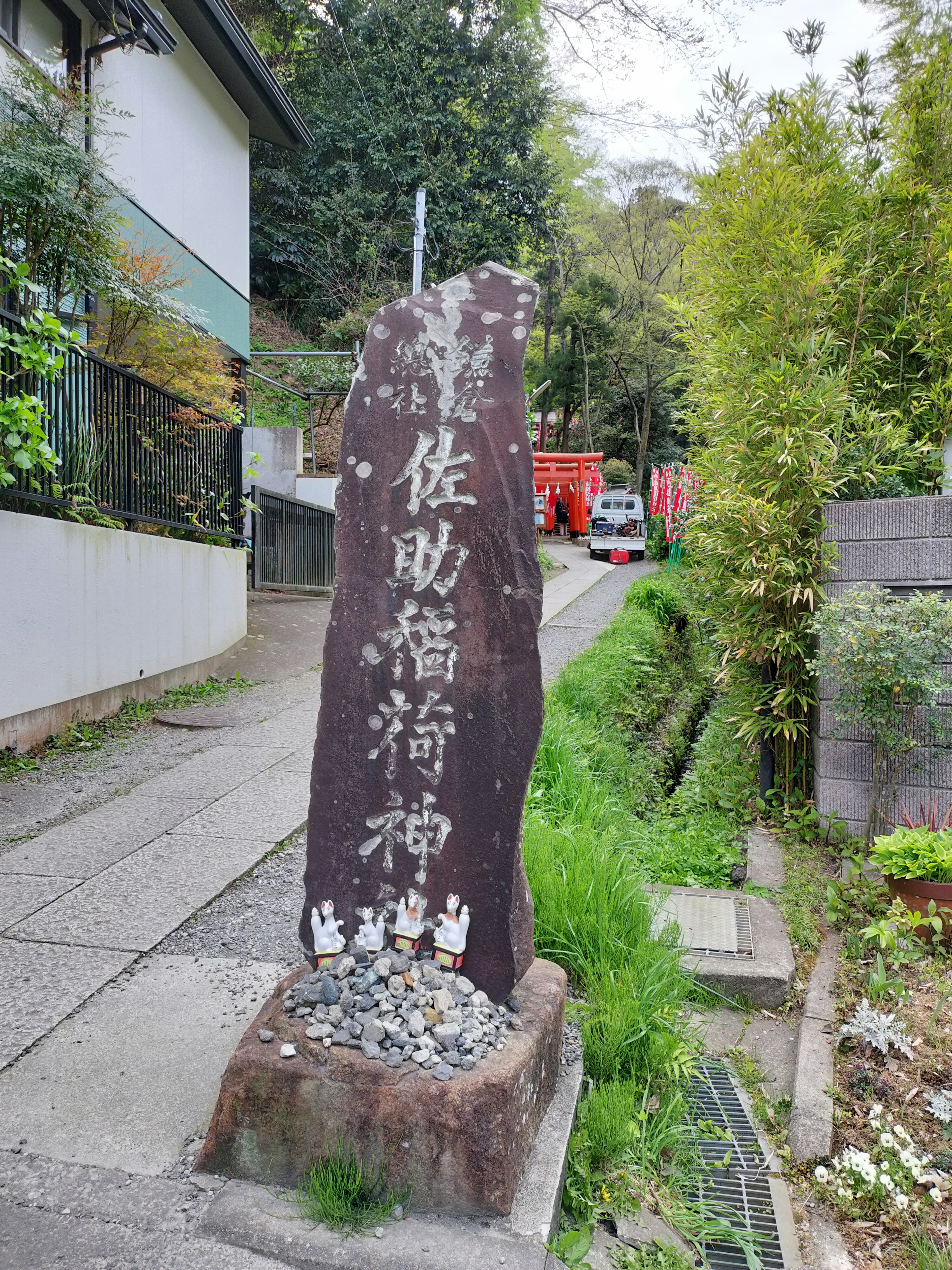 Un monumento de piedra rodeado de vegetación con un edificio blanco y una puerta torii roja al fondo