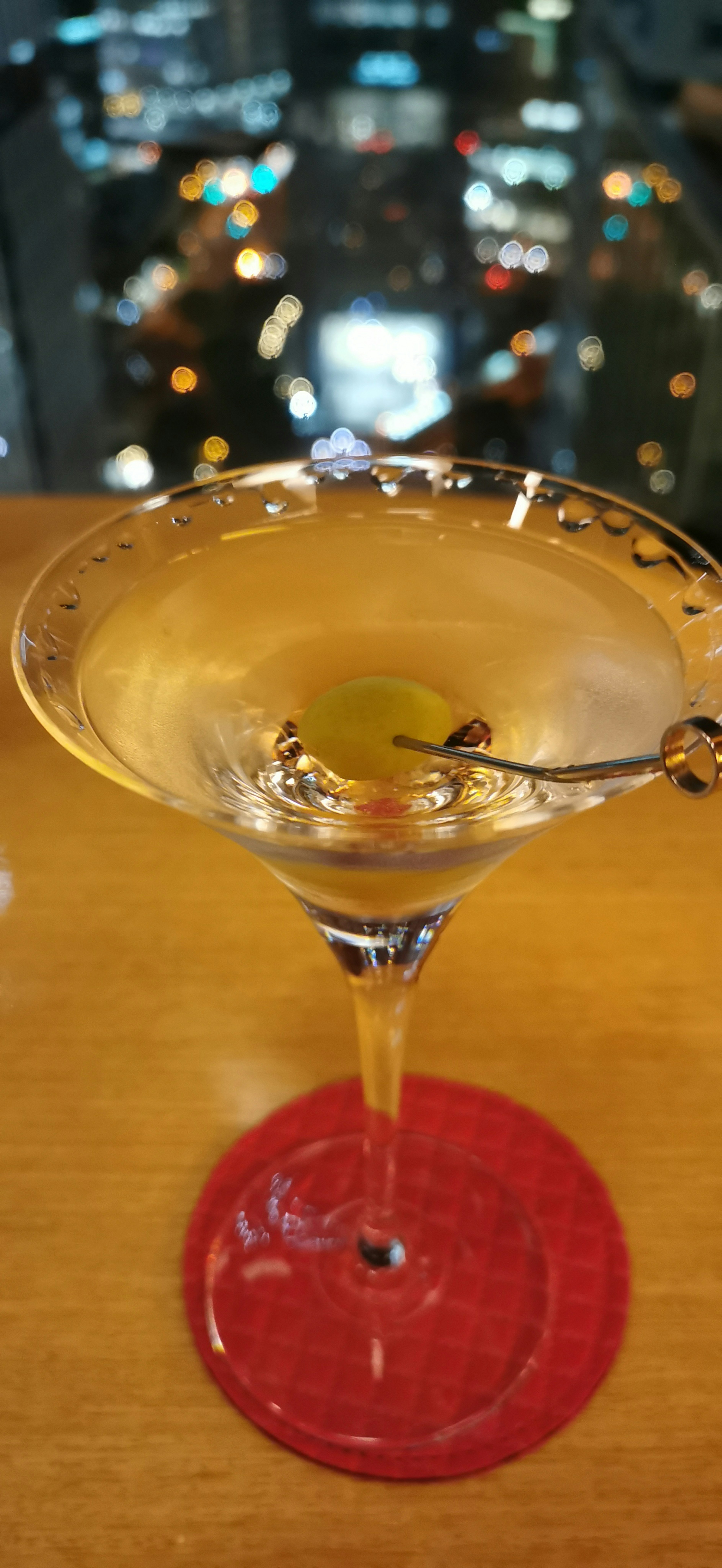 Cocktail glass with drink and olive on a red coaster overlooking a city night view