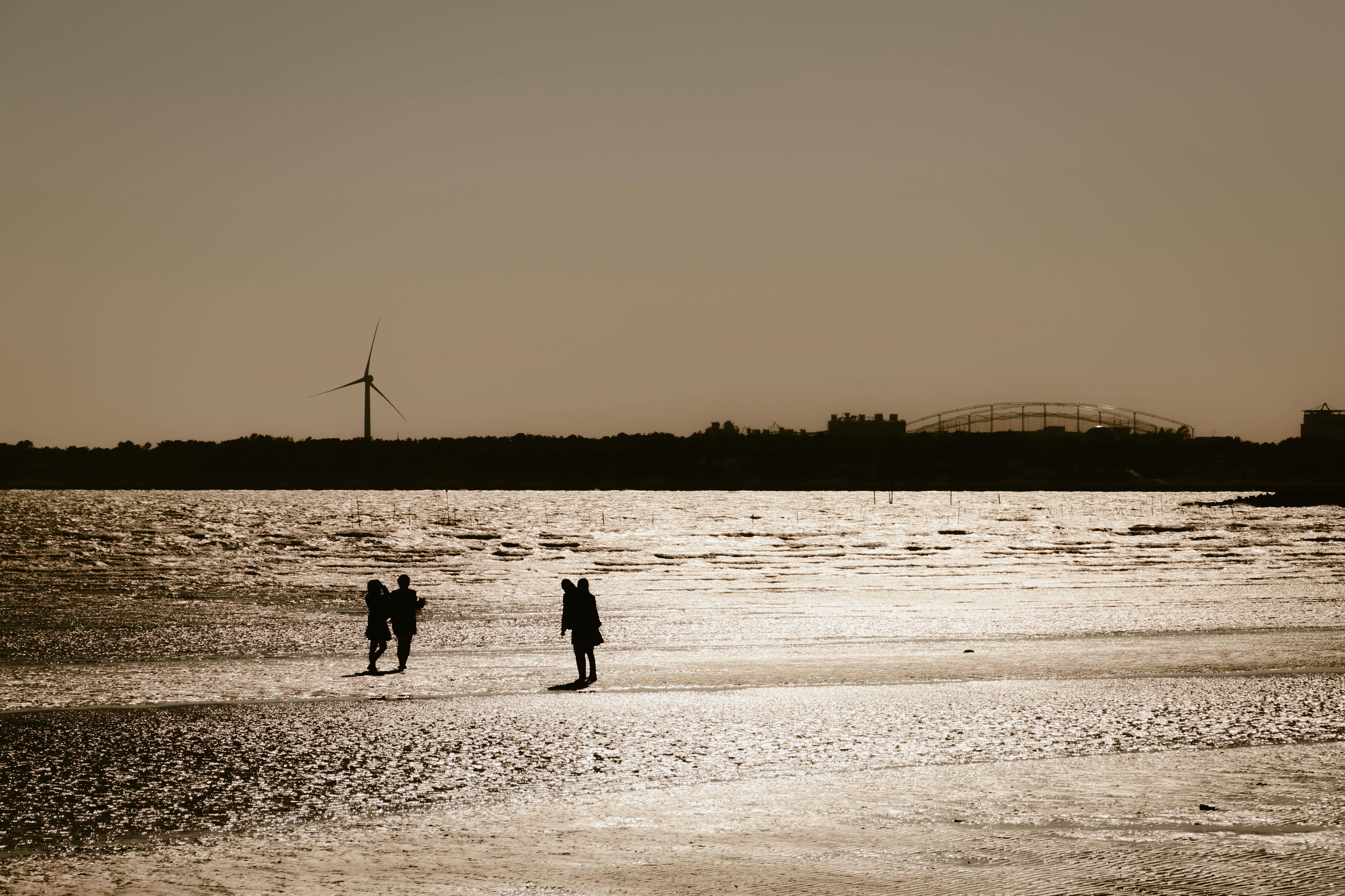 Siluetas de dos personas caminando en una playa al atardecer con un aerogenerador al fondo