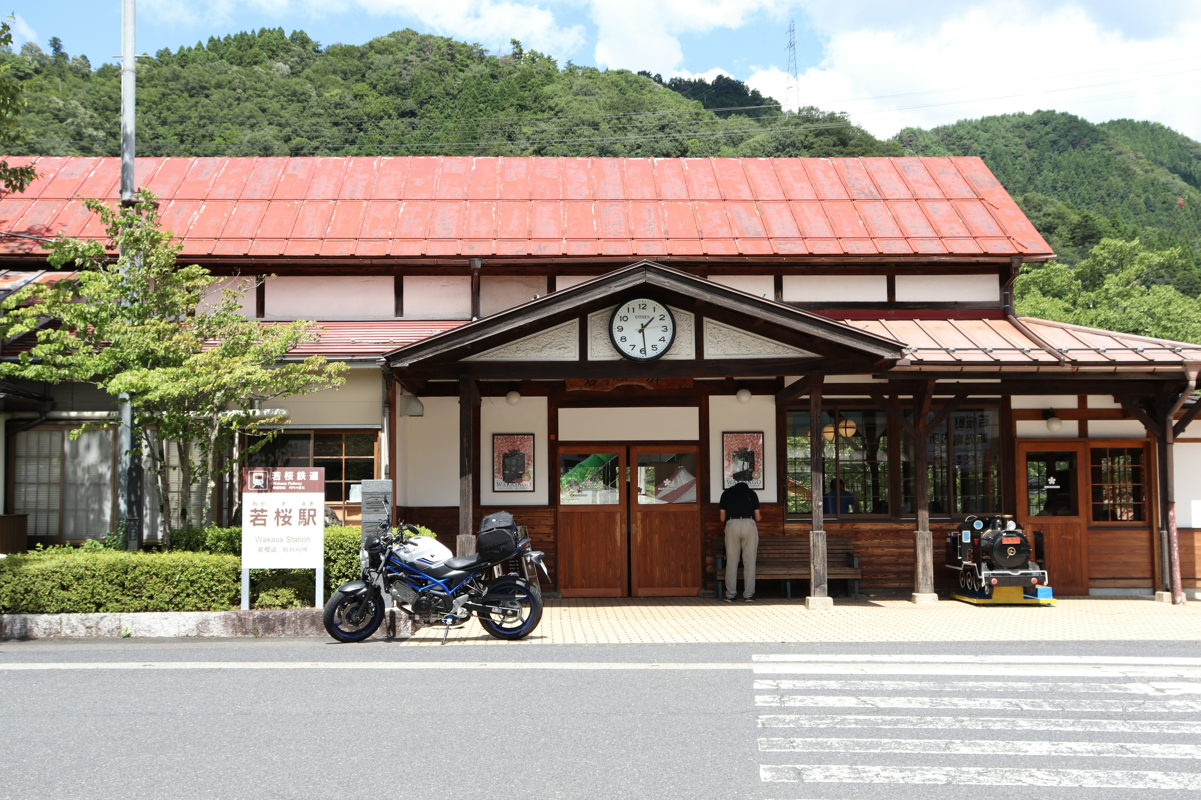 Traditional wooden train station with a red roof surrounded by green mountains and a motorcycle parked in front