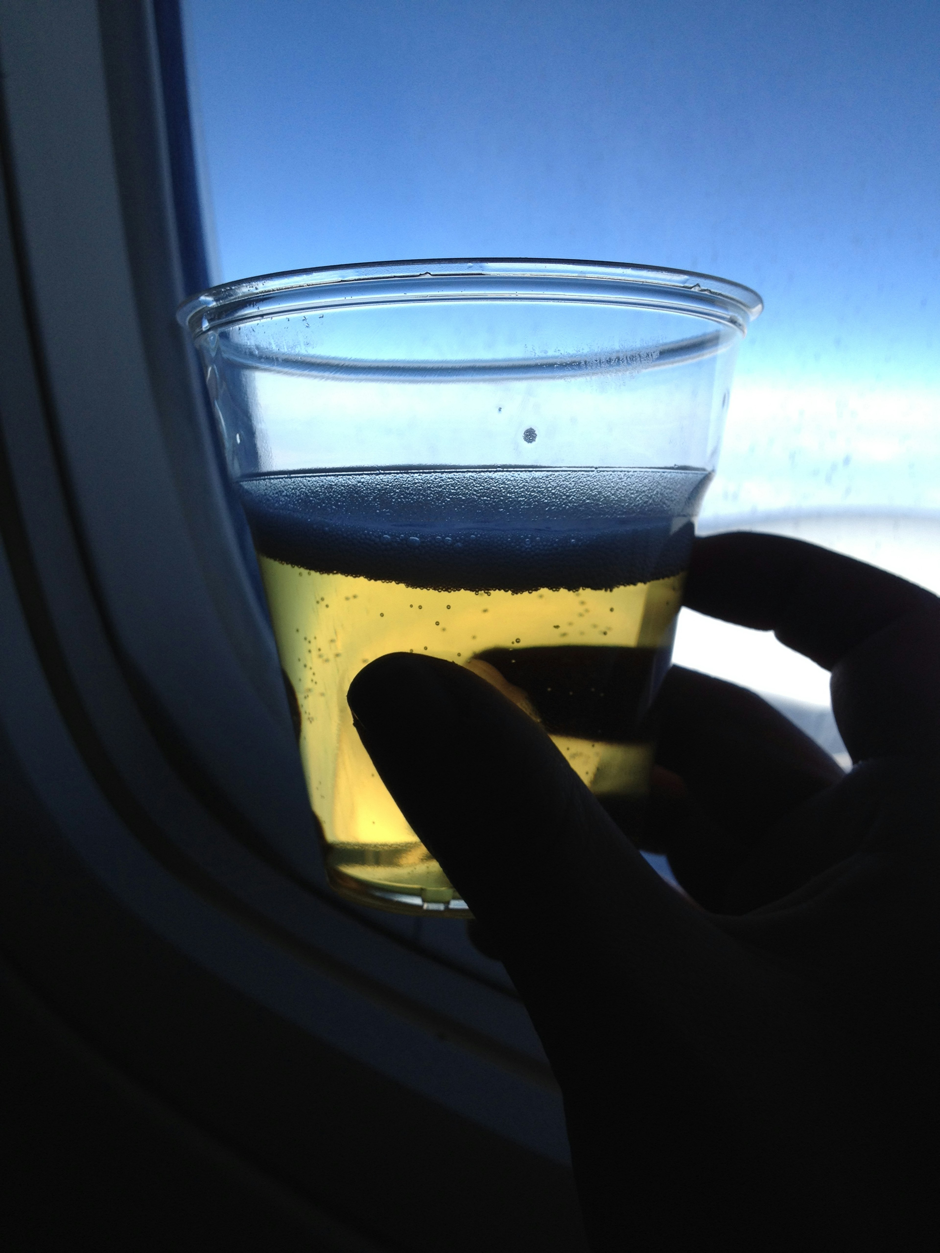 A hand holding a cup of beer against the backdrop of blue sky and clouds seen through an airplane window