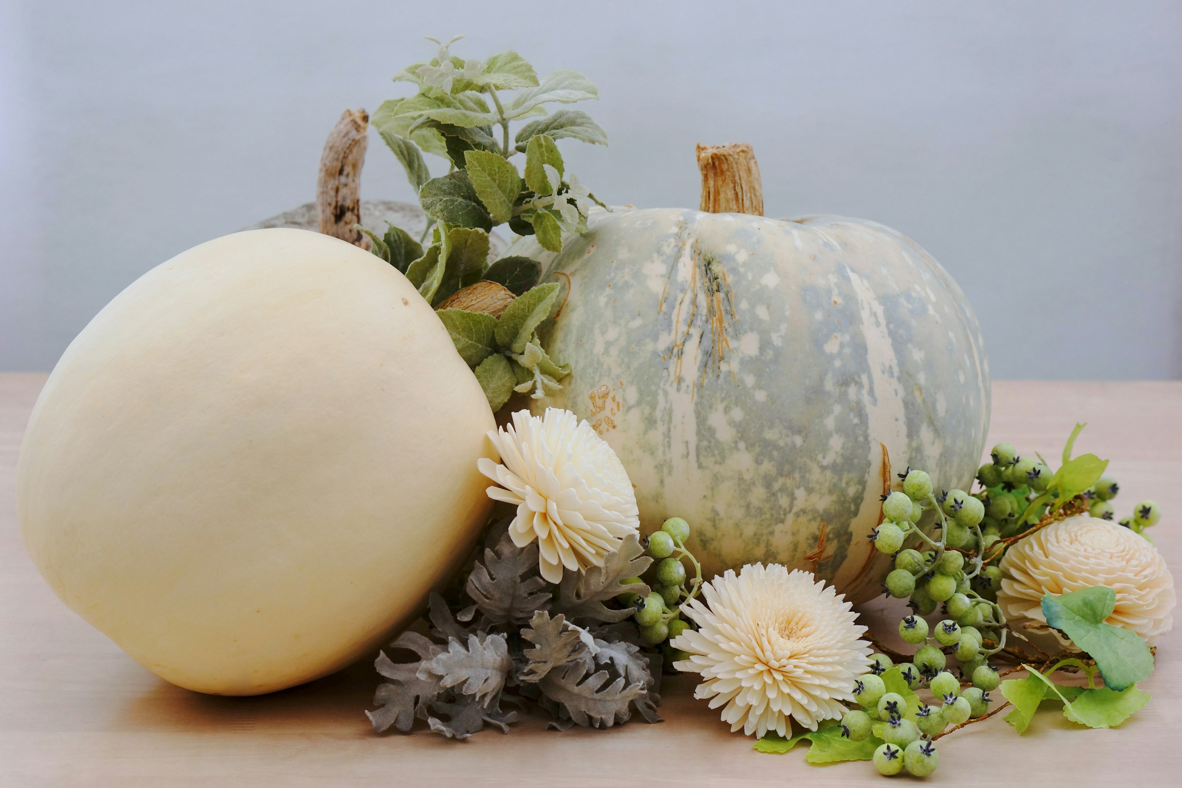 Una calabaza blanca y una calabaza verde dispuestas con plantas y flores alrededor