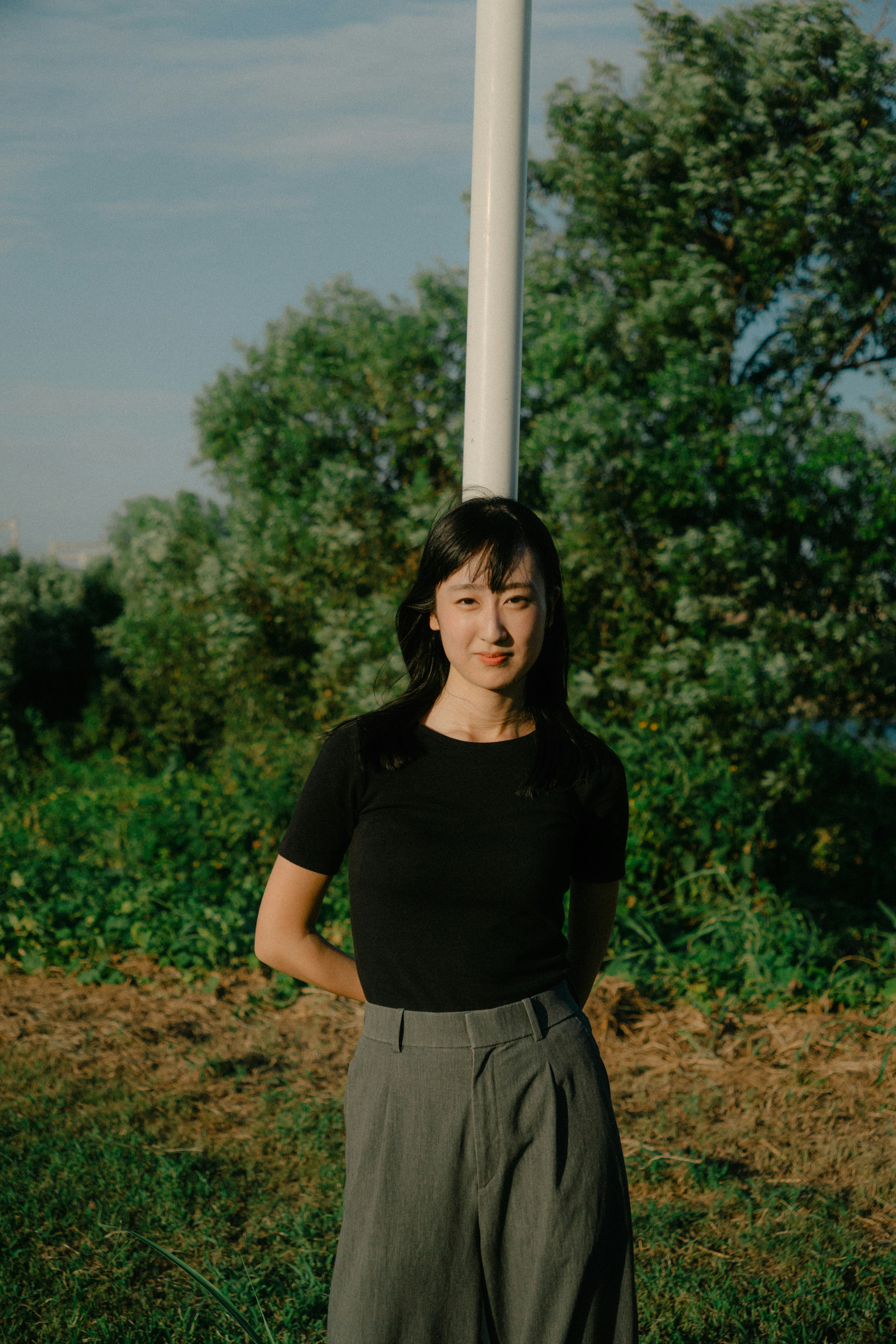 A woman stands in front of a green background wearing a black top and gray pants