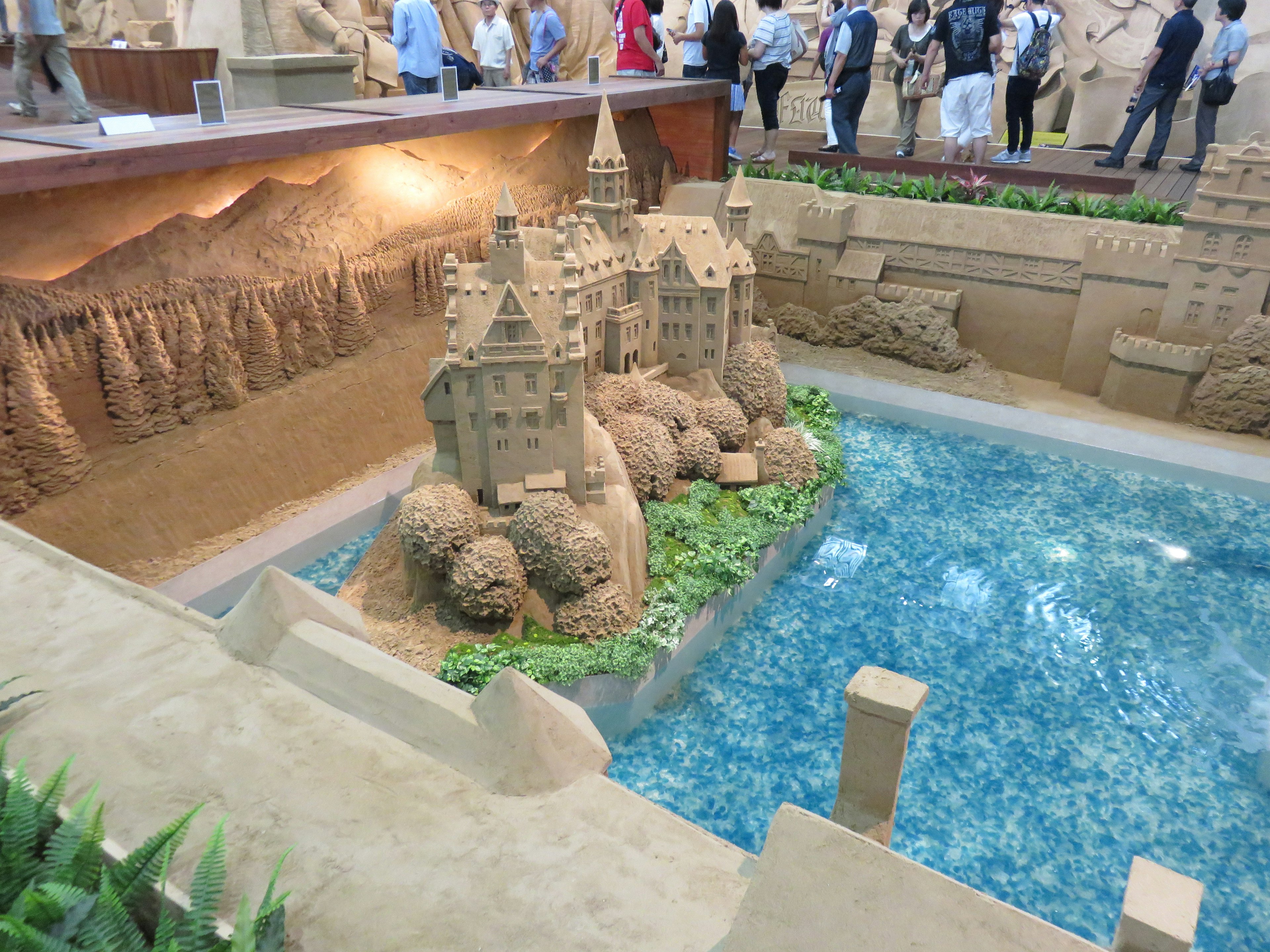 Intricate sand sculpture of a castle surrounded by a pond and greenery with visitors