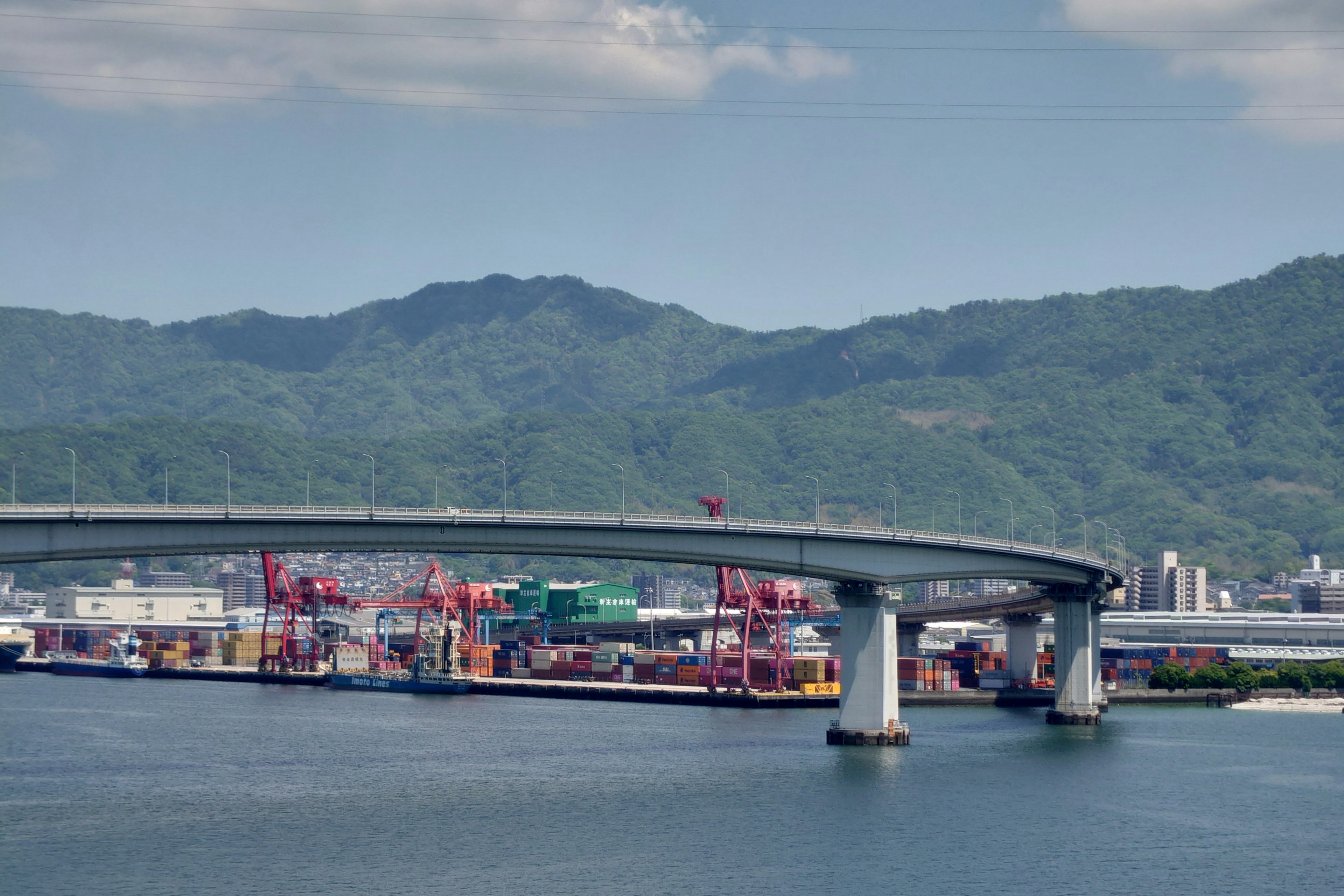 Malersicher Hafenblick mit blauem Meer und einer Brücke mit Containerschiffen und Bergen im Hintergrund