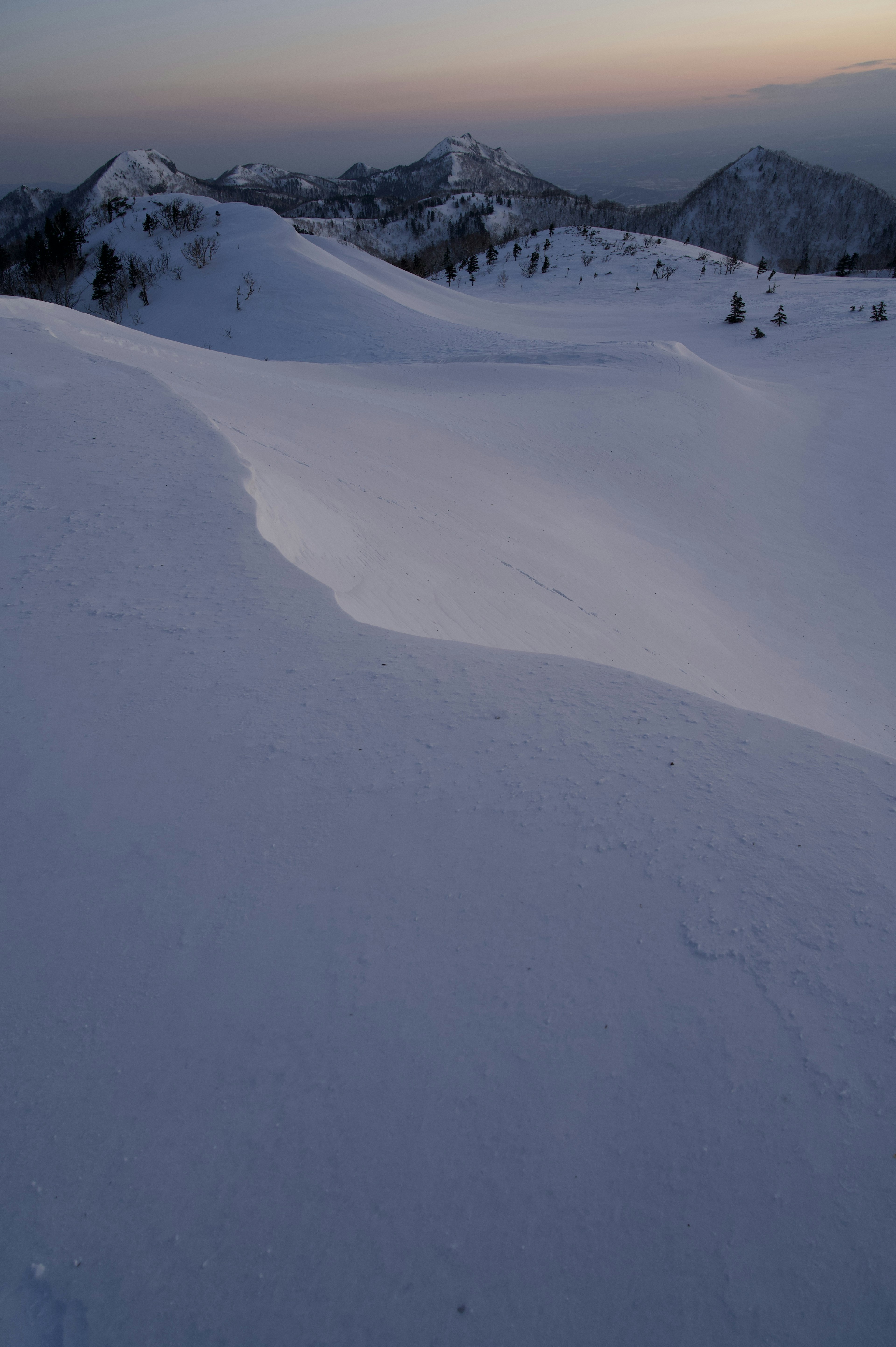 Paisaje montañoso cubierto de nieve con suaves curvas y cielo crepuscular