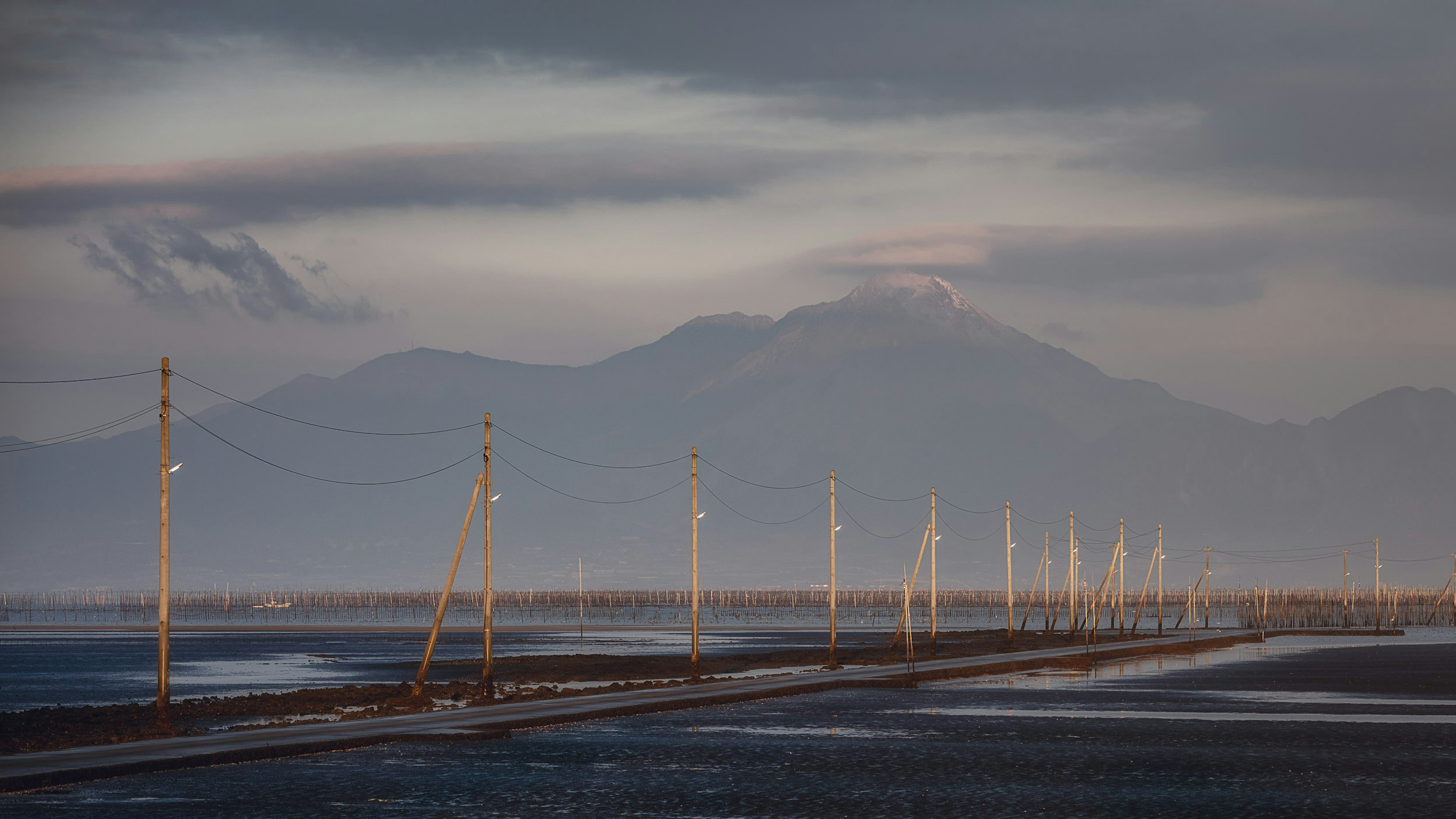 穏やかな空の下に並ぶ電柱と遠くの山々が広がる風景