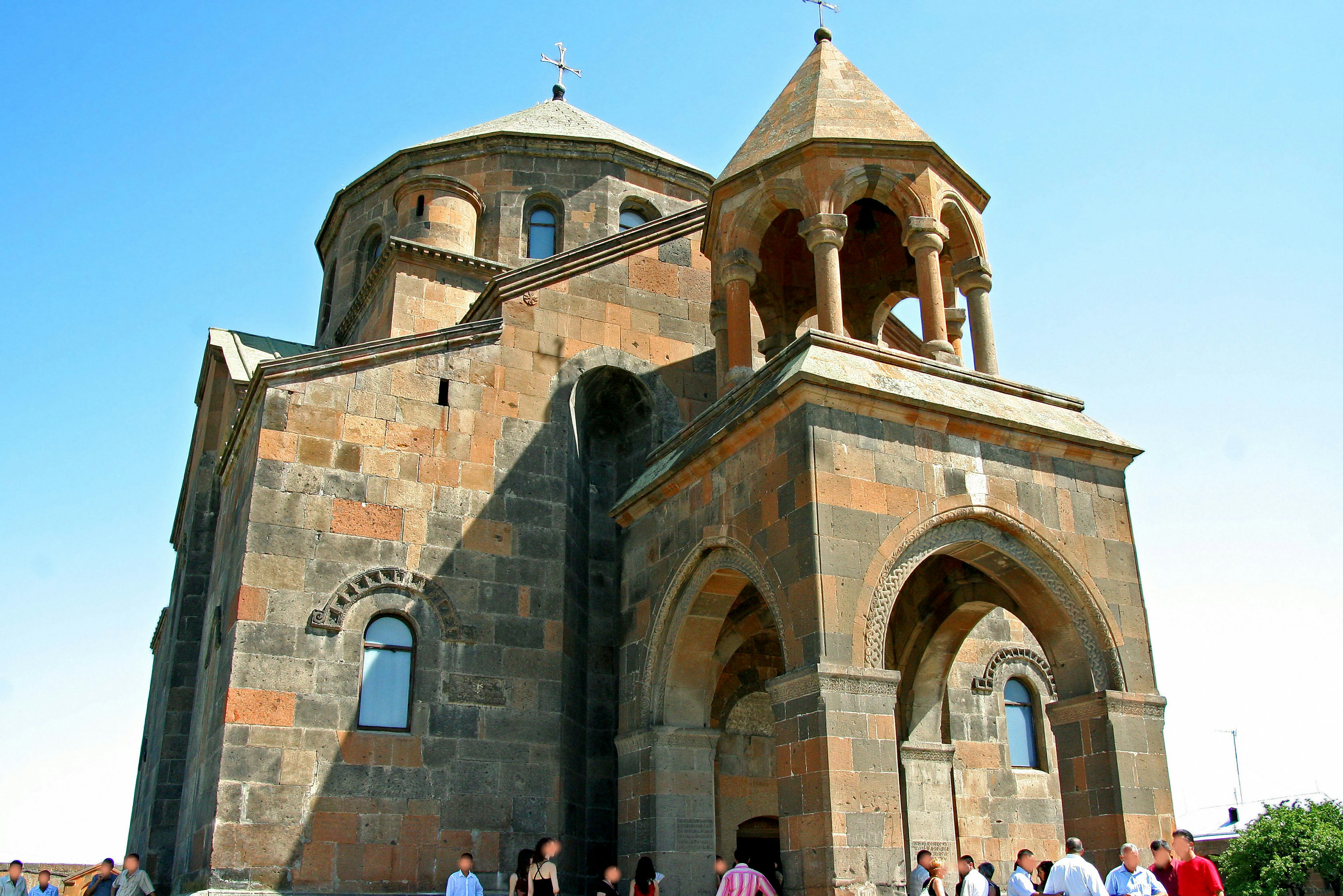 Bild einer armenischen Kirche, die ihre markante Architektur unter einem klaren blauen Himmel mit Besuchern zeigt