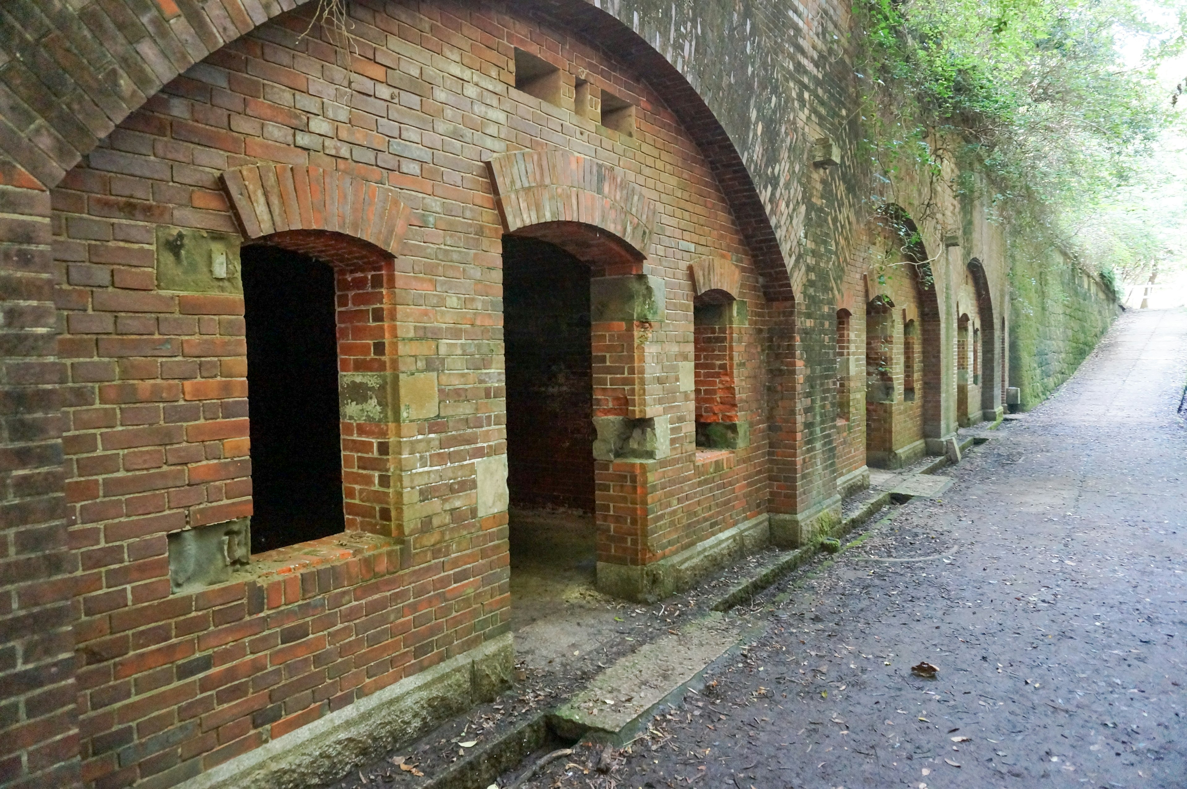 Vue latérale d'un ancien bâtiment en briques avec des fenêtres en arc et une végétation luxuriante