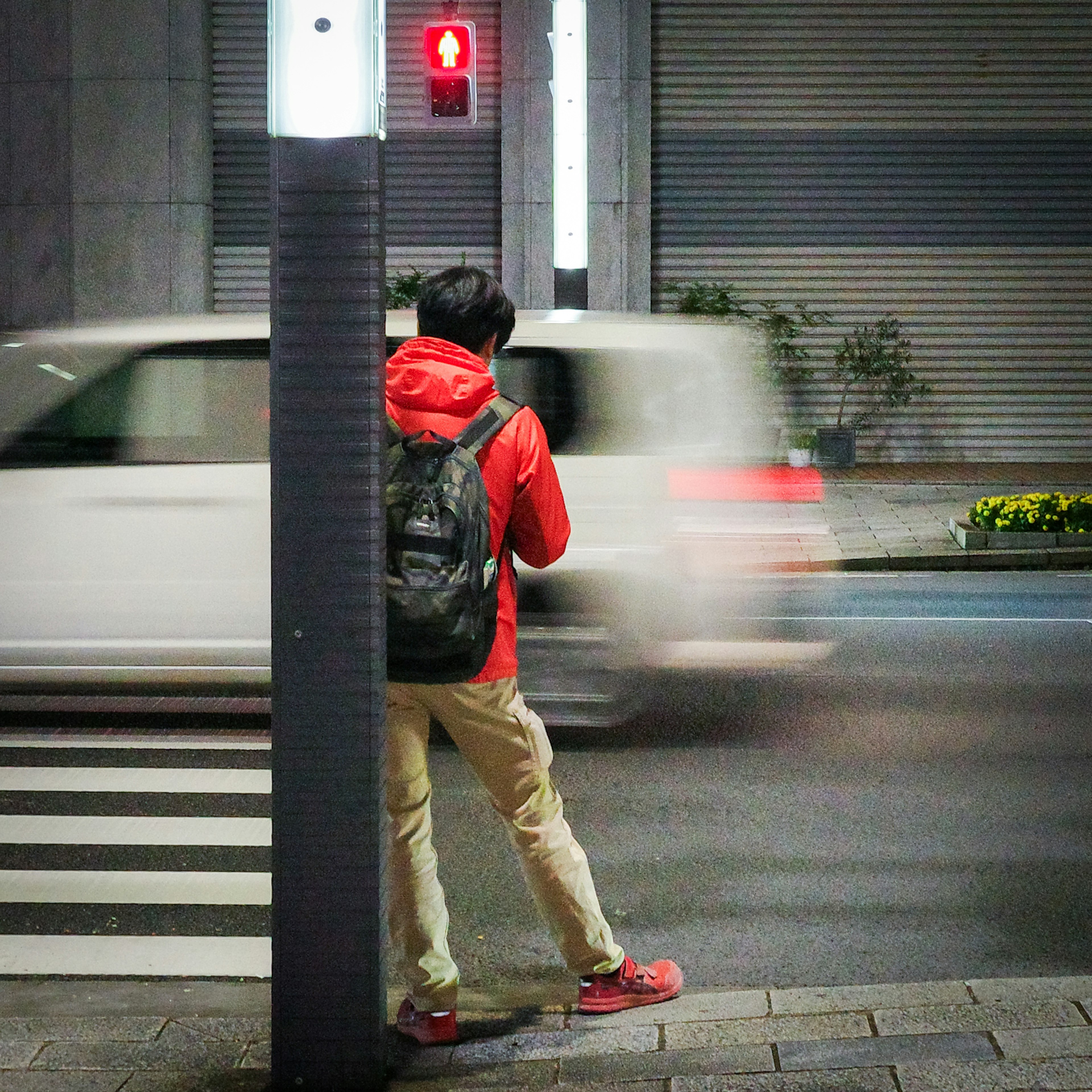 A person in a red jacket waiting at a traffic light in a city setting