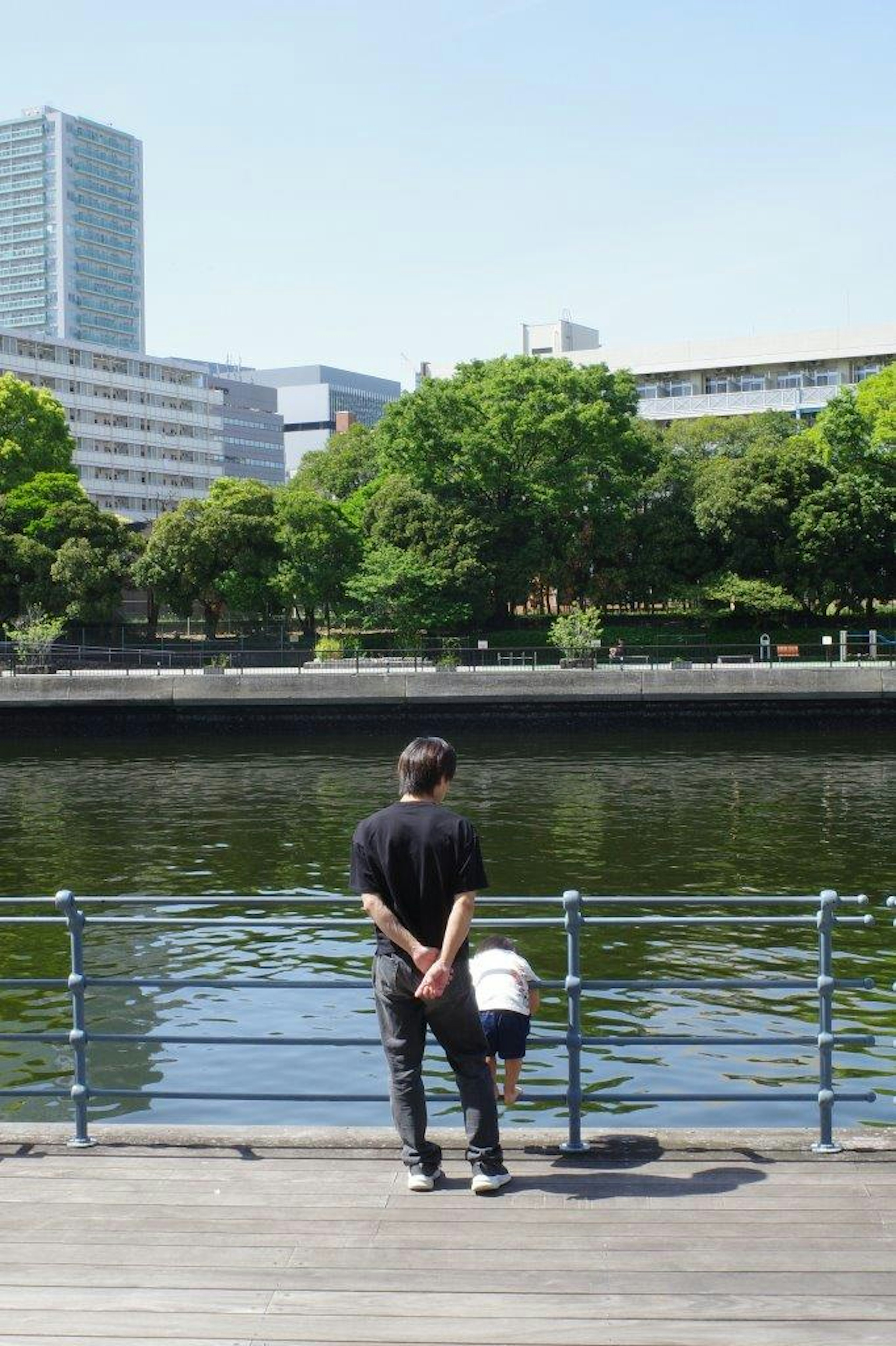 Un hombre y un niño de pie junto a un río tranquilo mirando el paisaje urbano con vegetación