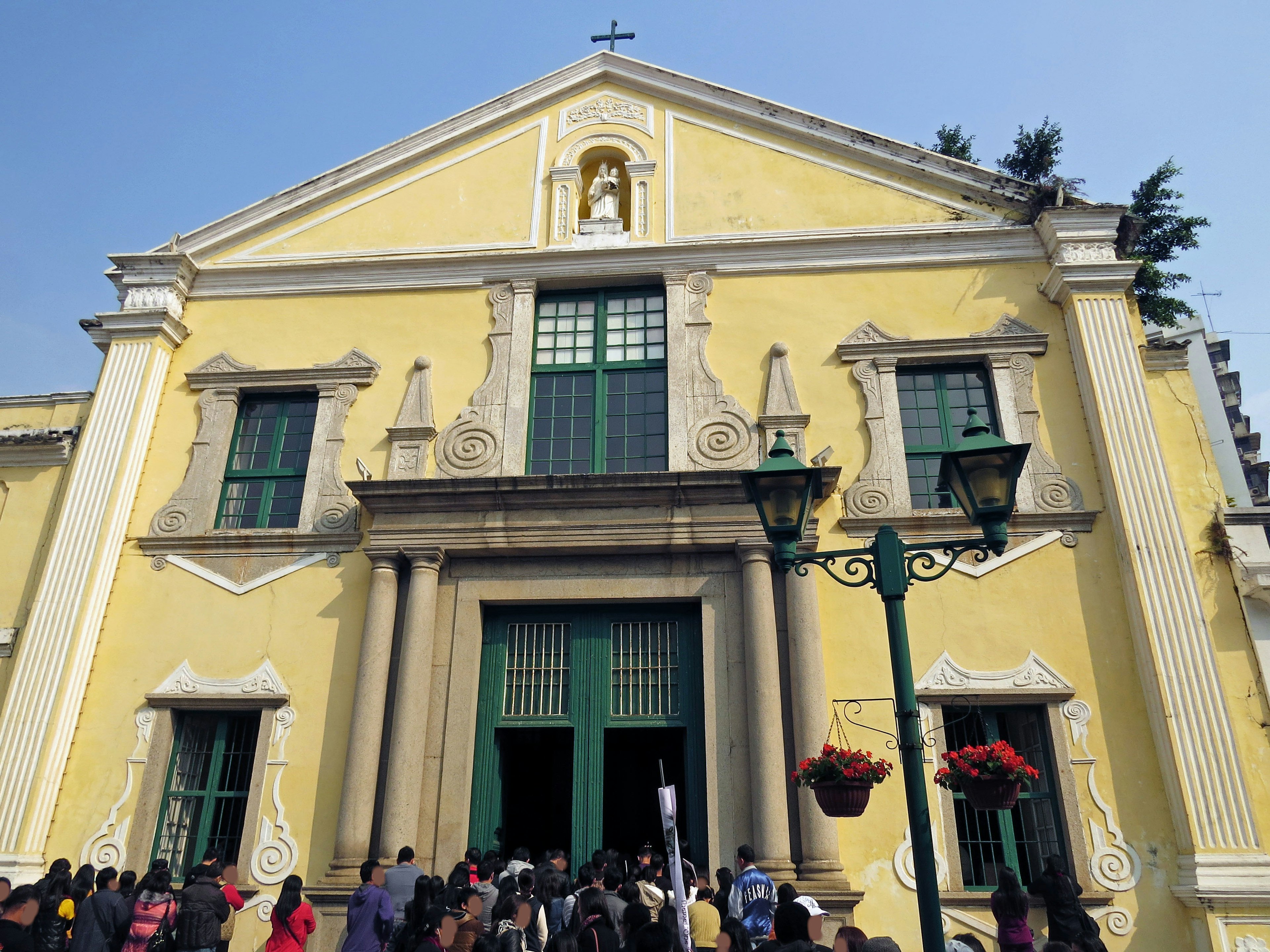 Persone si riuniscono davanti a un edificio giallo con finestre verdi e una grande porta, con una scultura di croce in cima