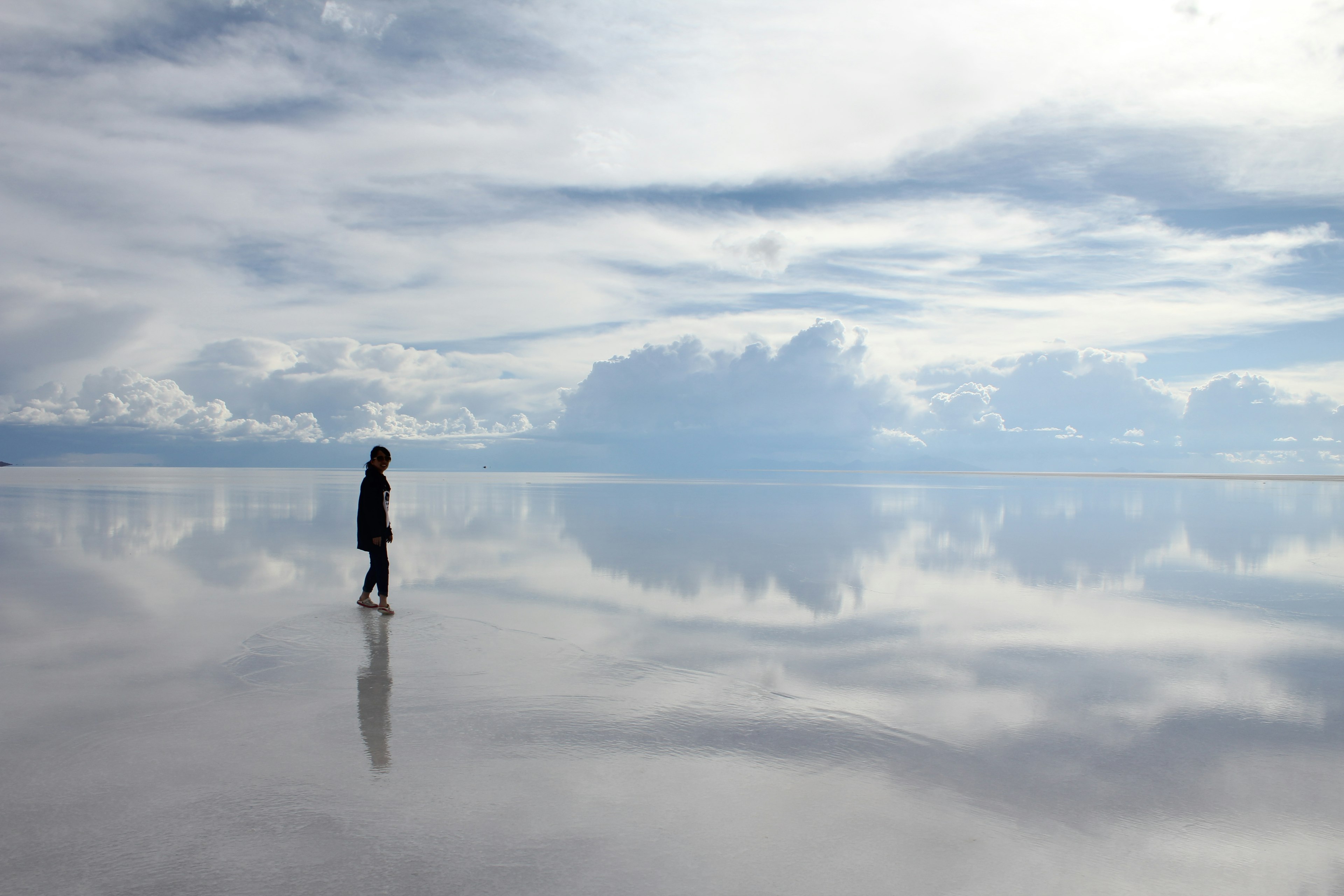 A person standing on a salt flat reflecting the blue sky and white clouds