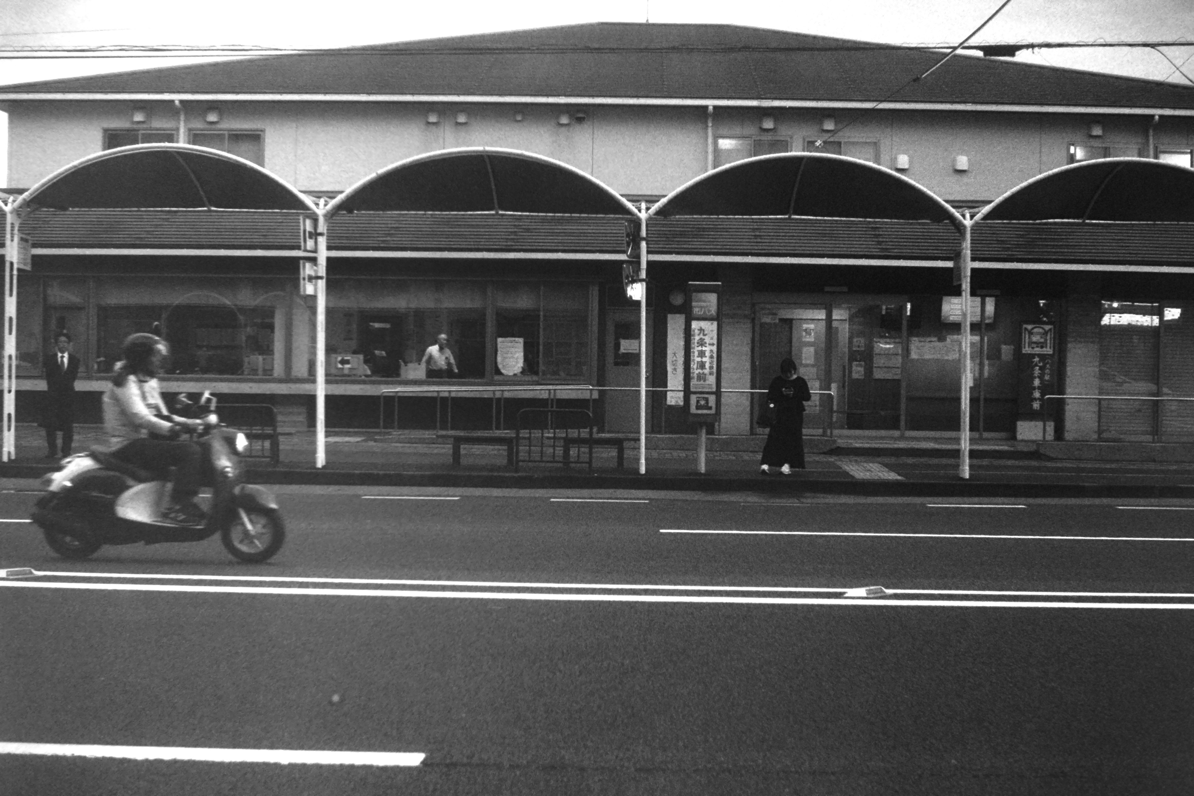 Scène de rue en noir et blanc avec un scooter passant devant des boutiques à arcade traditionnelles