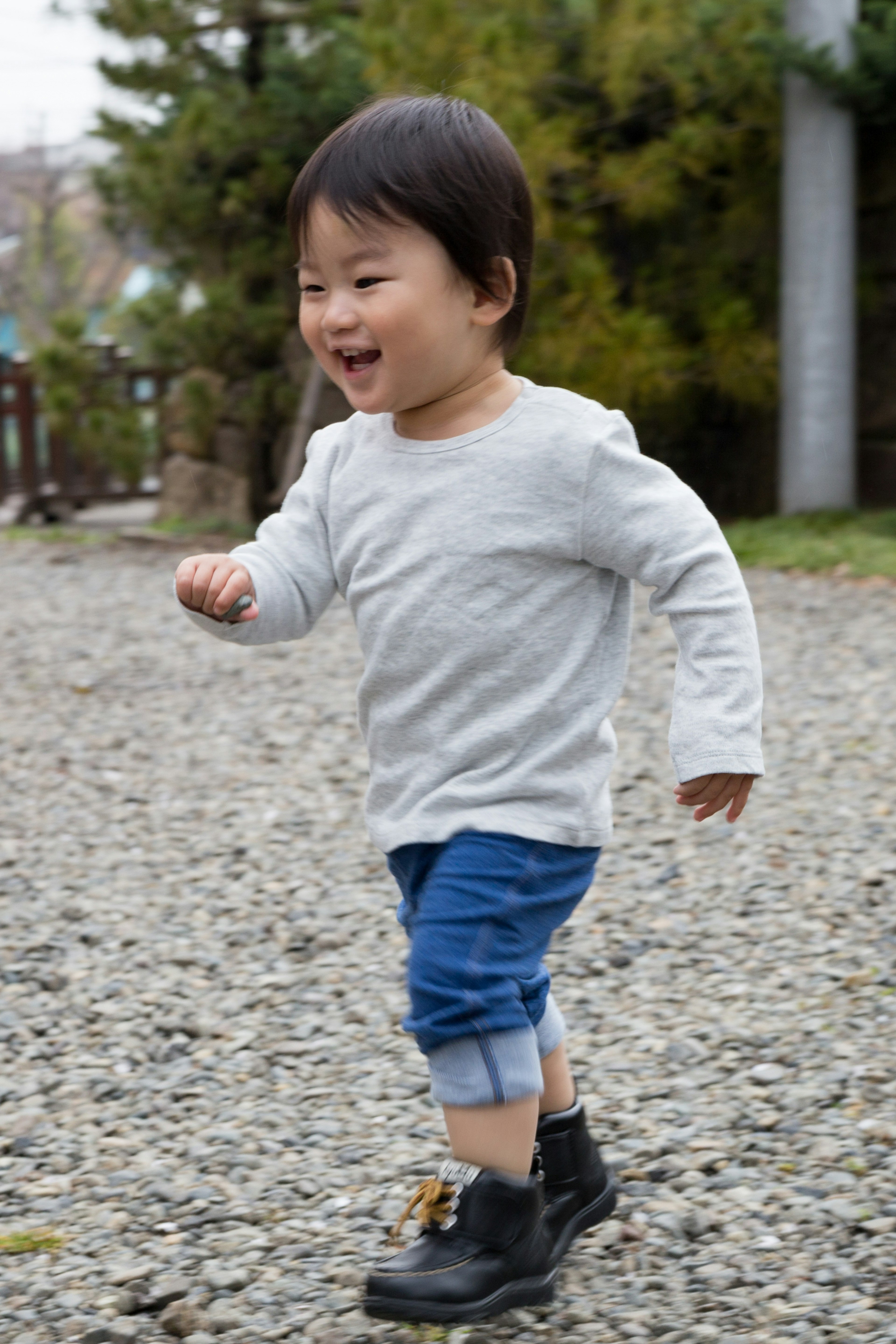 Happy young boy running wearing gray long-sleeve shirt and blue pants with black boots on a gravel path