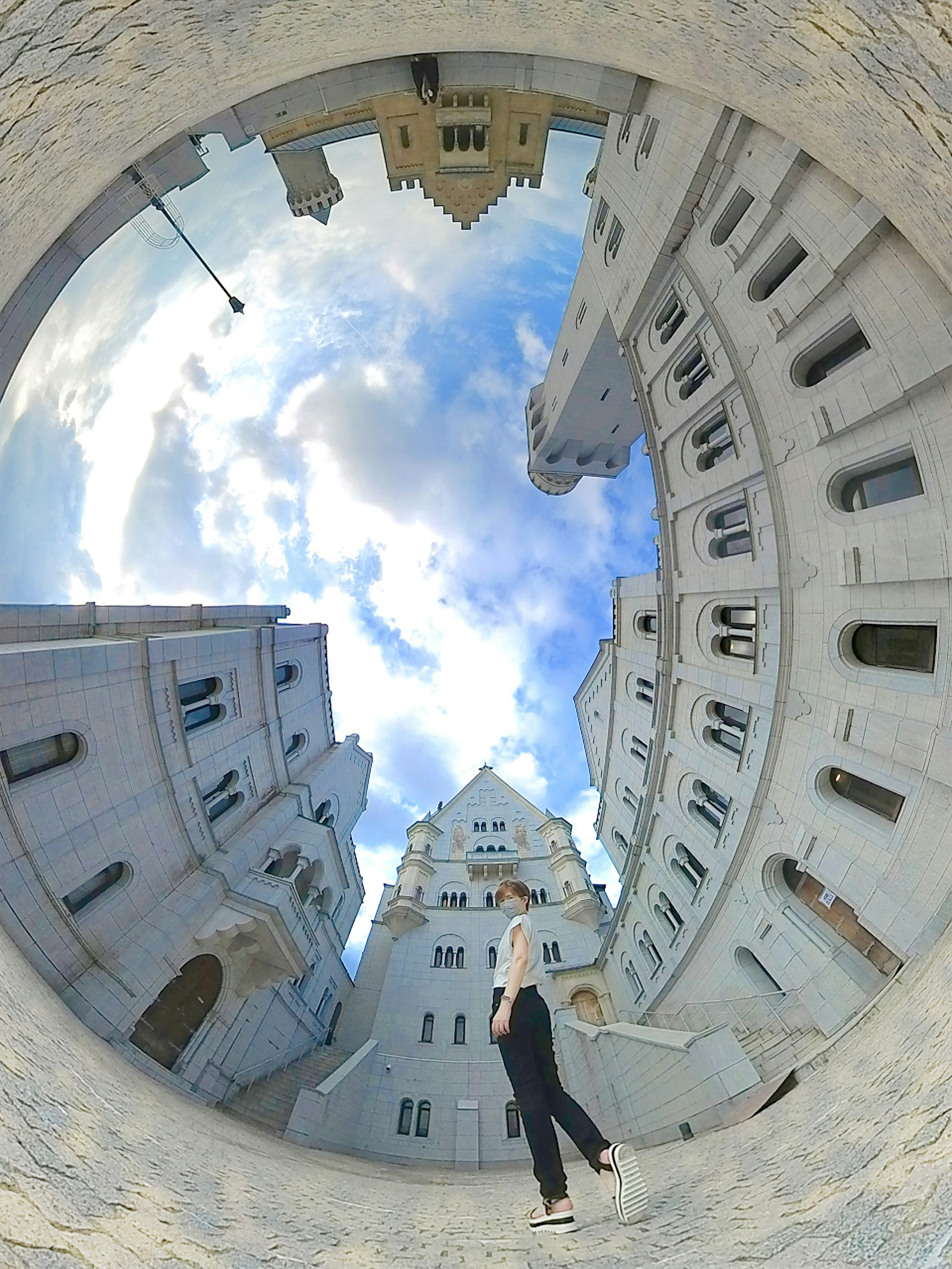 Person standing in a circular courtyard surrounded by white buildings with a blue sky and clouds above