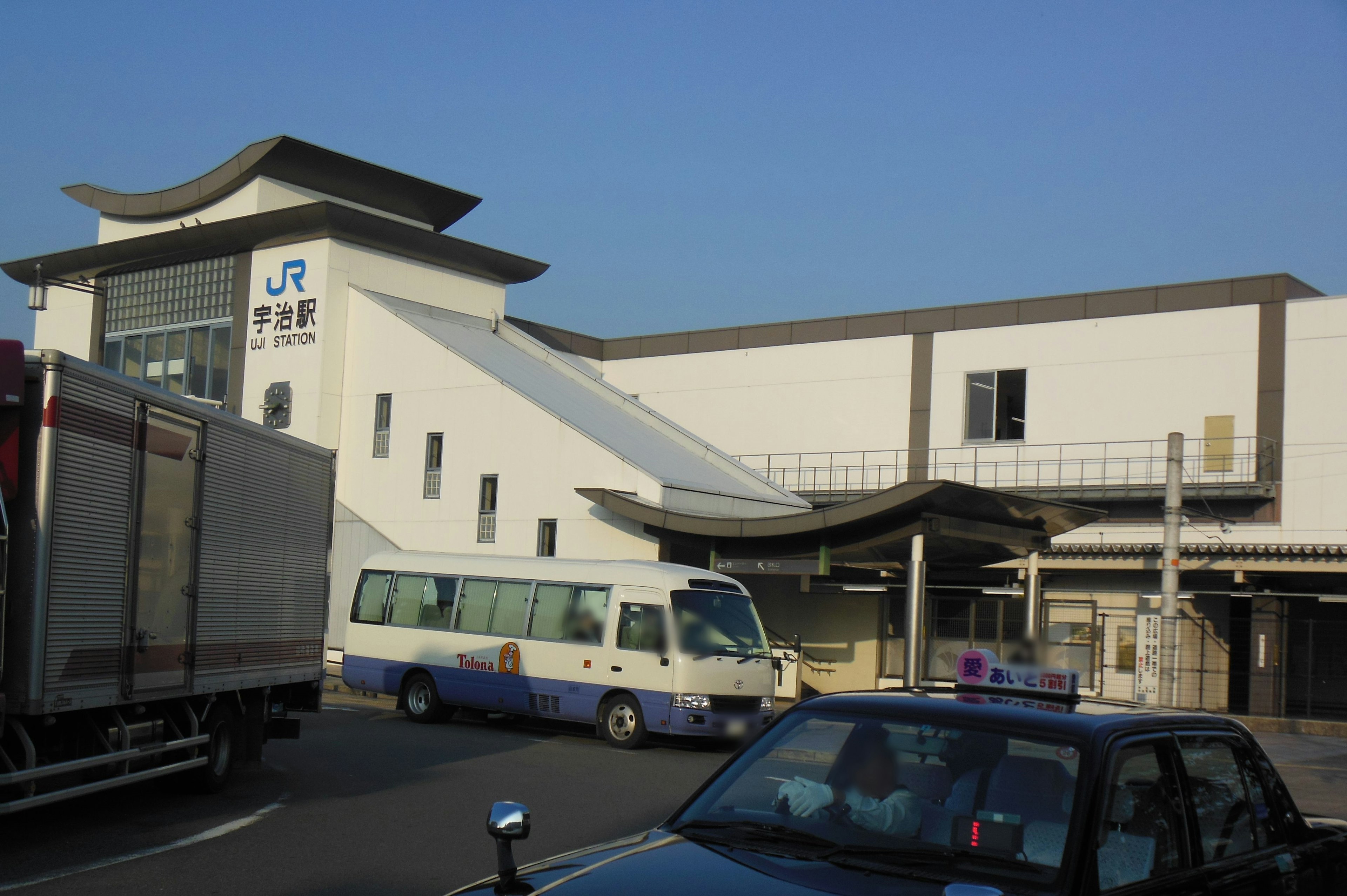 Vue d'une gare JR avec un bâtiment blanc sous un ciel bleu présentant un bus et des voitures garées