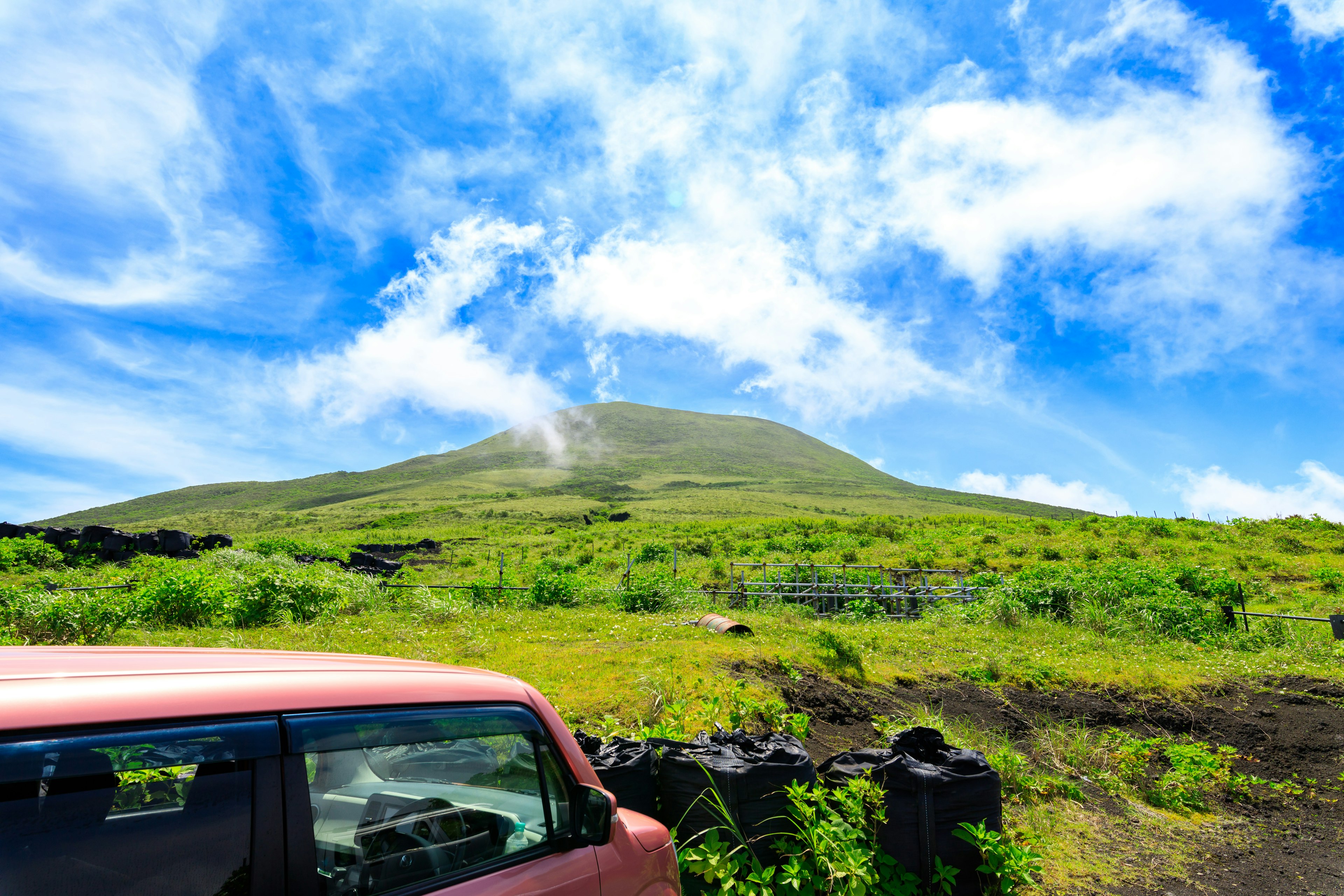 Una montaña rodeada de praderas verdes y un cielo azul con un coche rojo en primer plano