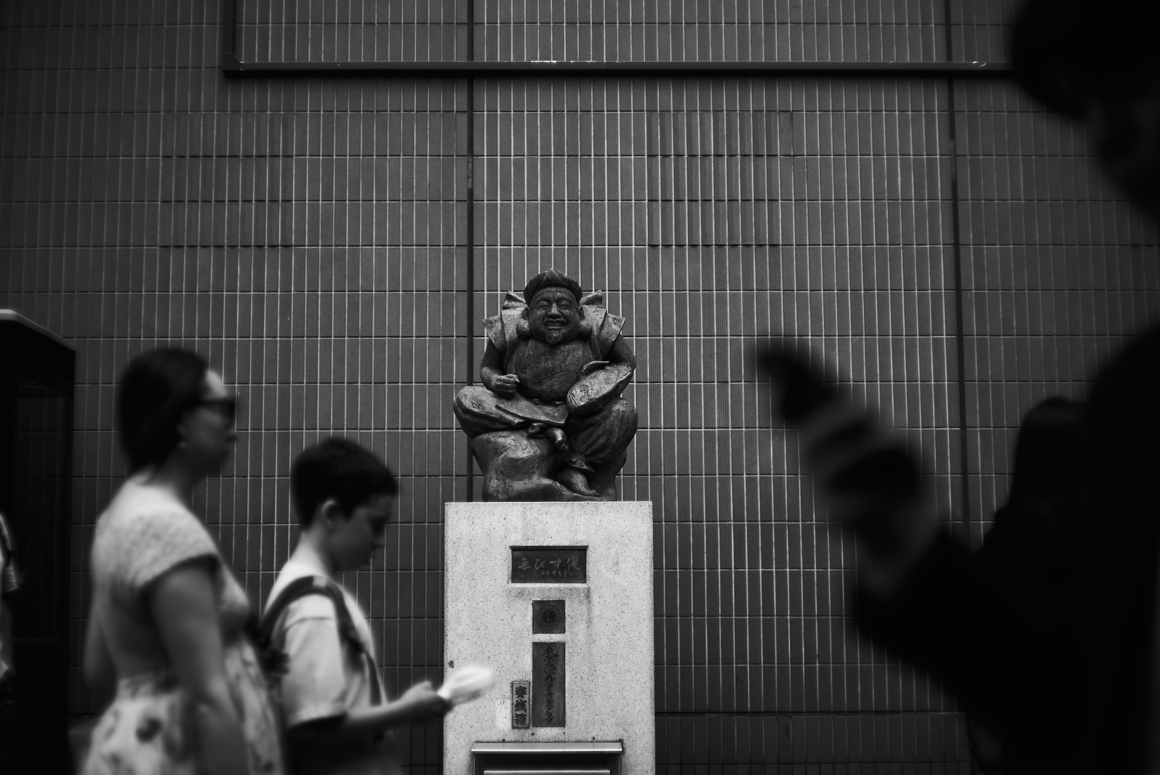 Una persona sentada en un pedestal en un entorno urbano en blanco y negro con transeúntes