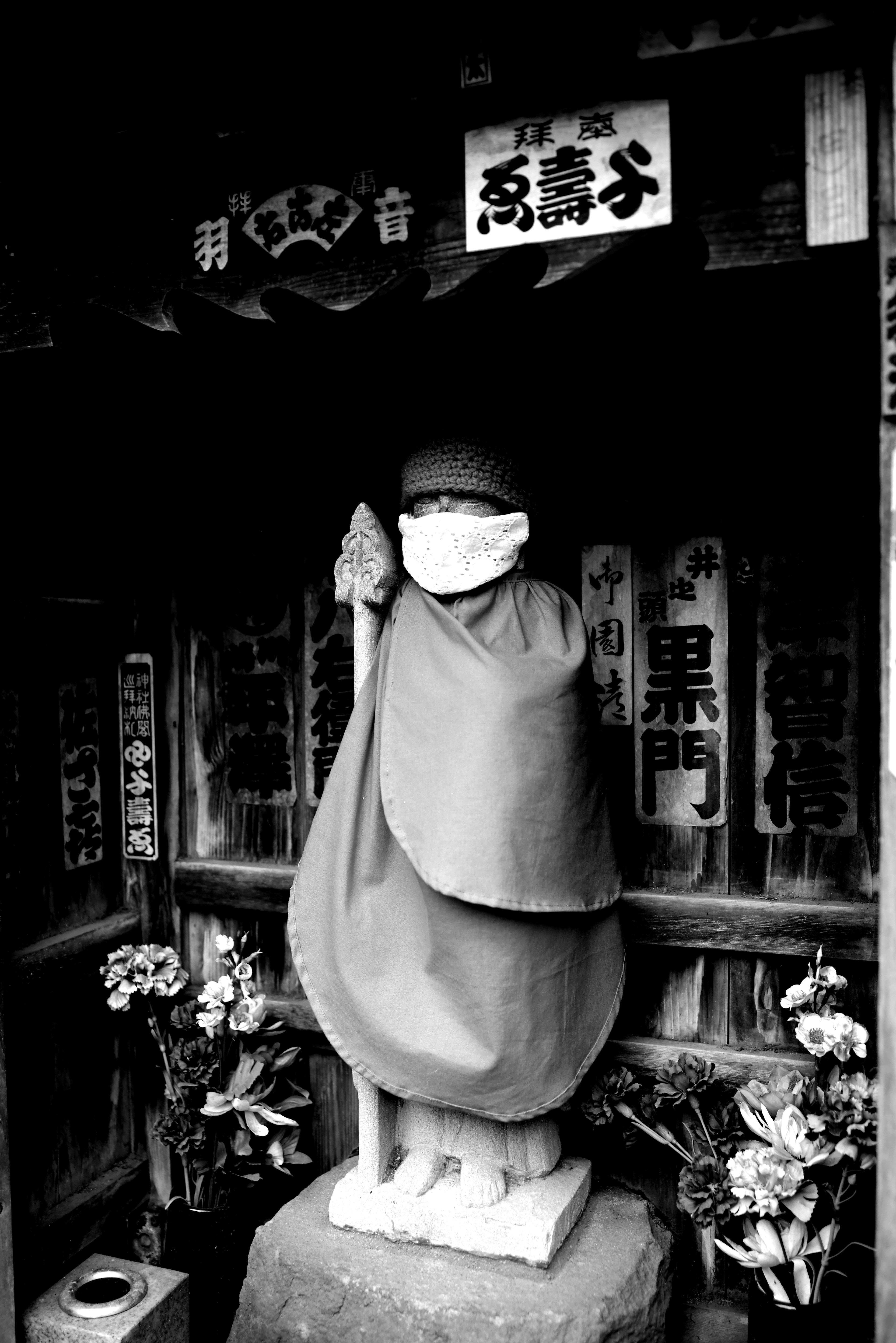 A black and white statue of a monk wearing a mask surrounded by flowers and old signage