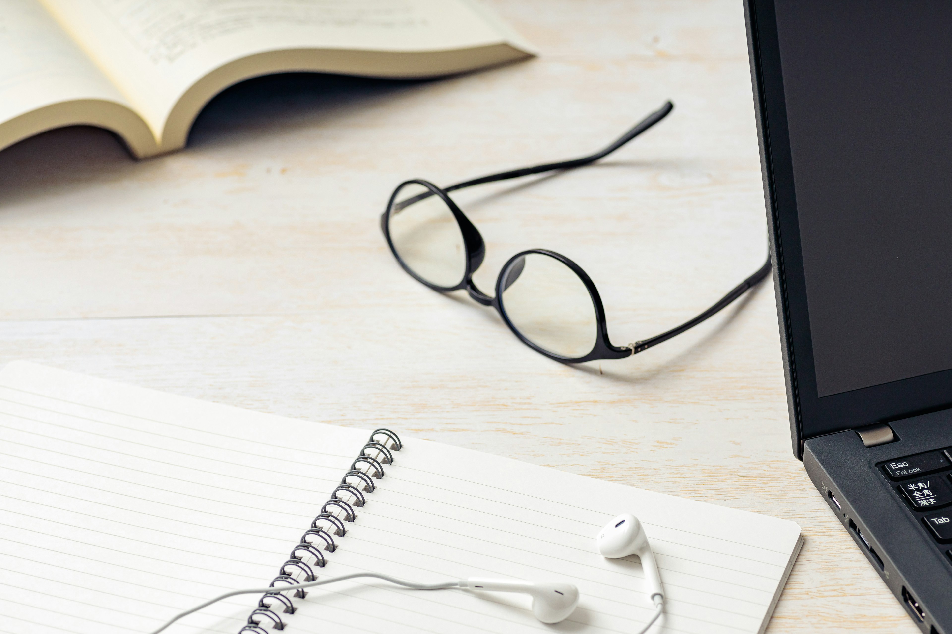 An open book, glasses, a laptop, a notebook, and earphones on a desk