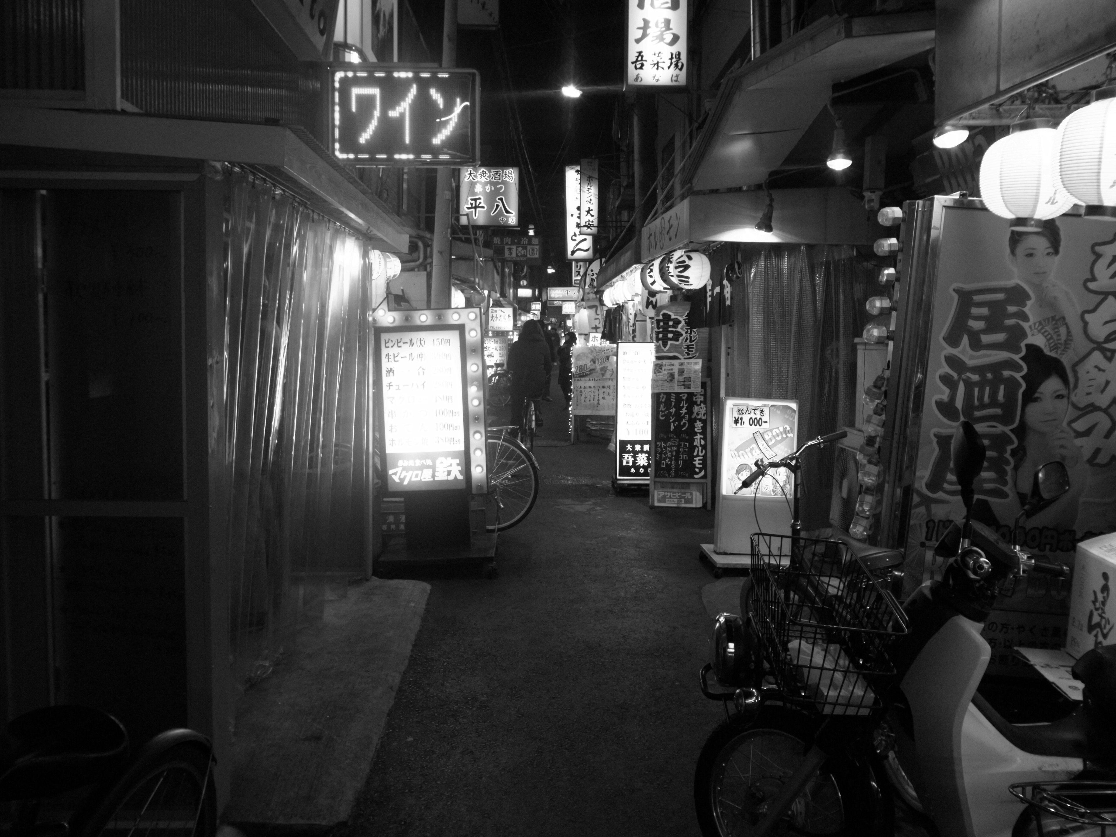 Foto en blanco y negro de un callejón estrecho en un distrito nocturno japonés con puestos de comida y letreros