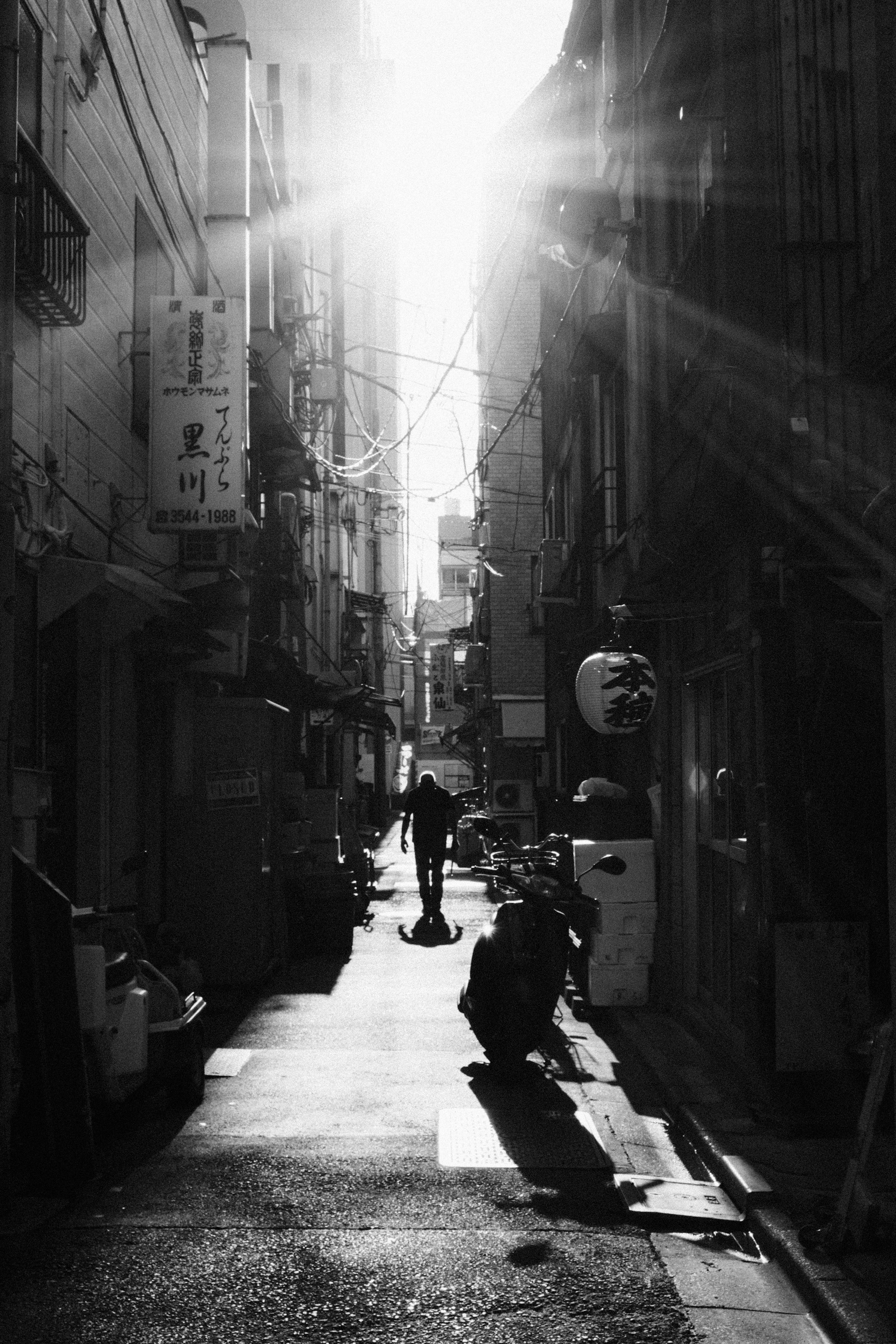 Black and white photo of a narrow alley with a person walking and bright light shining