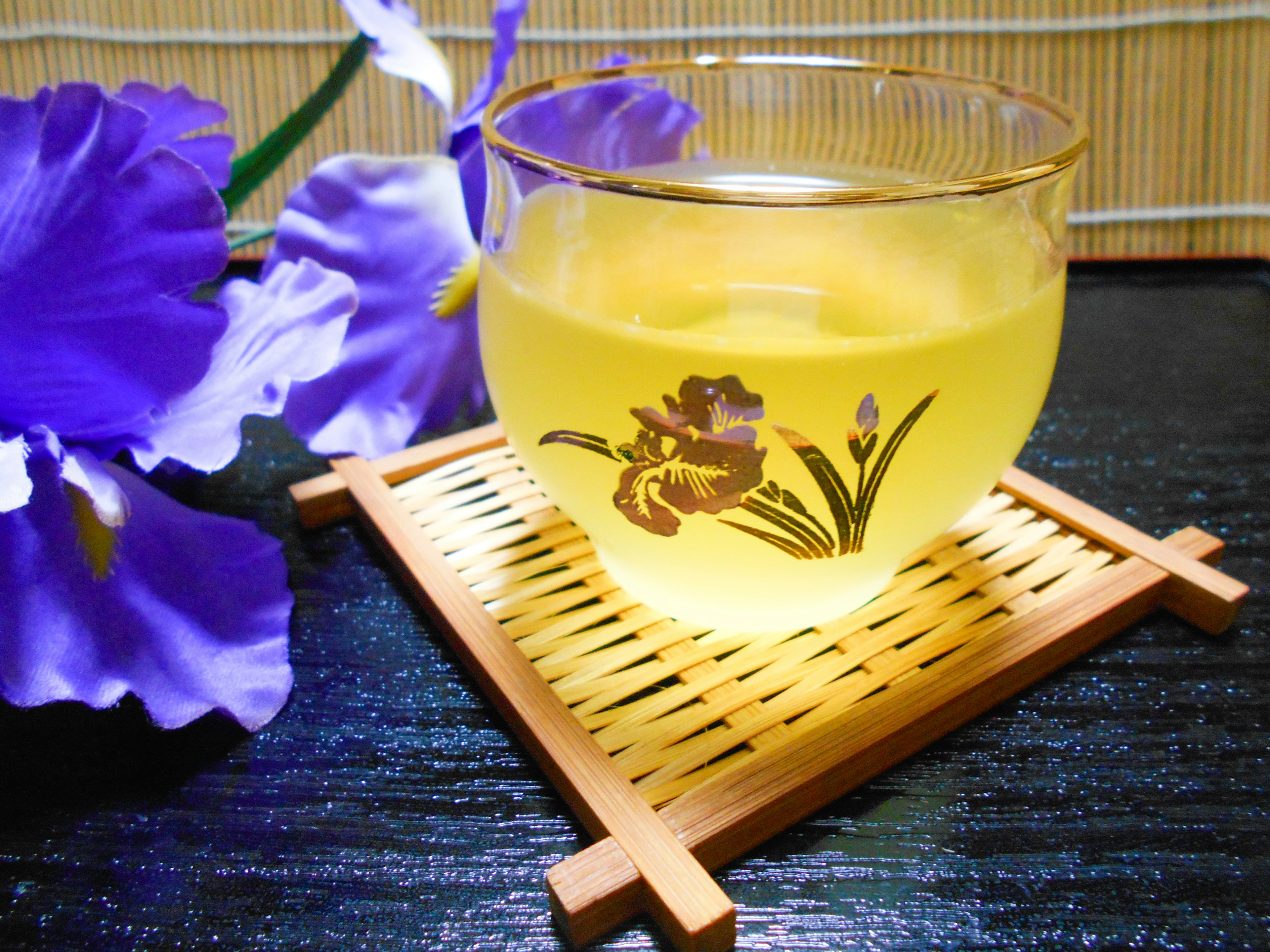 A glass with floral design filled with yellow drink Purple flowers in the background