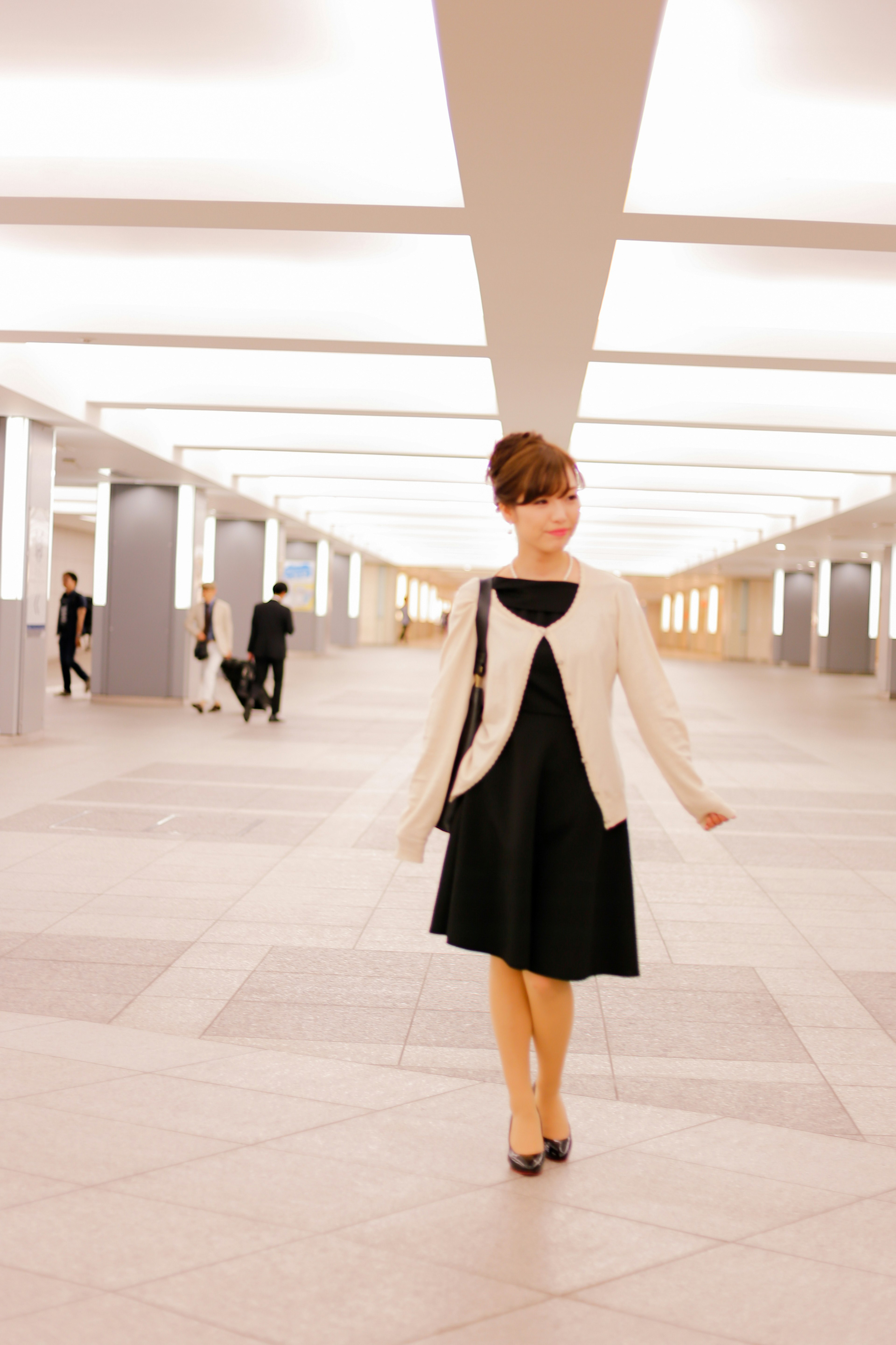 Une femme en robe noire et cardigan blanc marchant dans un couloir lumineux