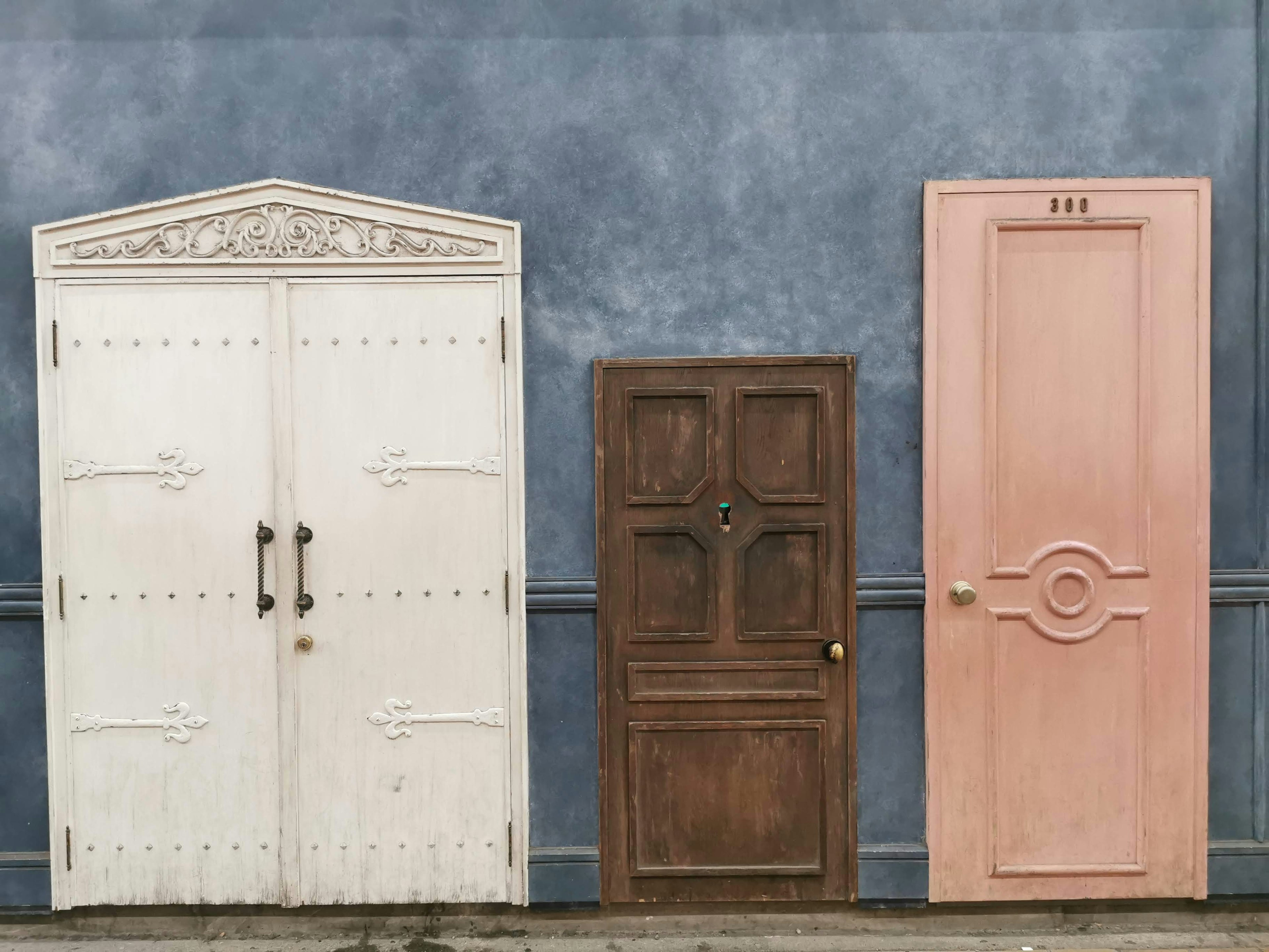 Tres puertas distintas alineadas contra una pared azul incluyendo una puerta decorativa blanca una puerta vintage marrón y una puerta rosa