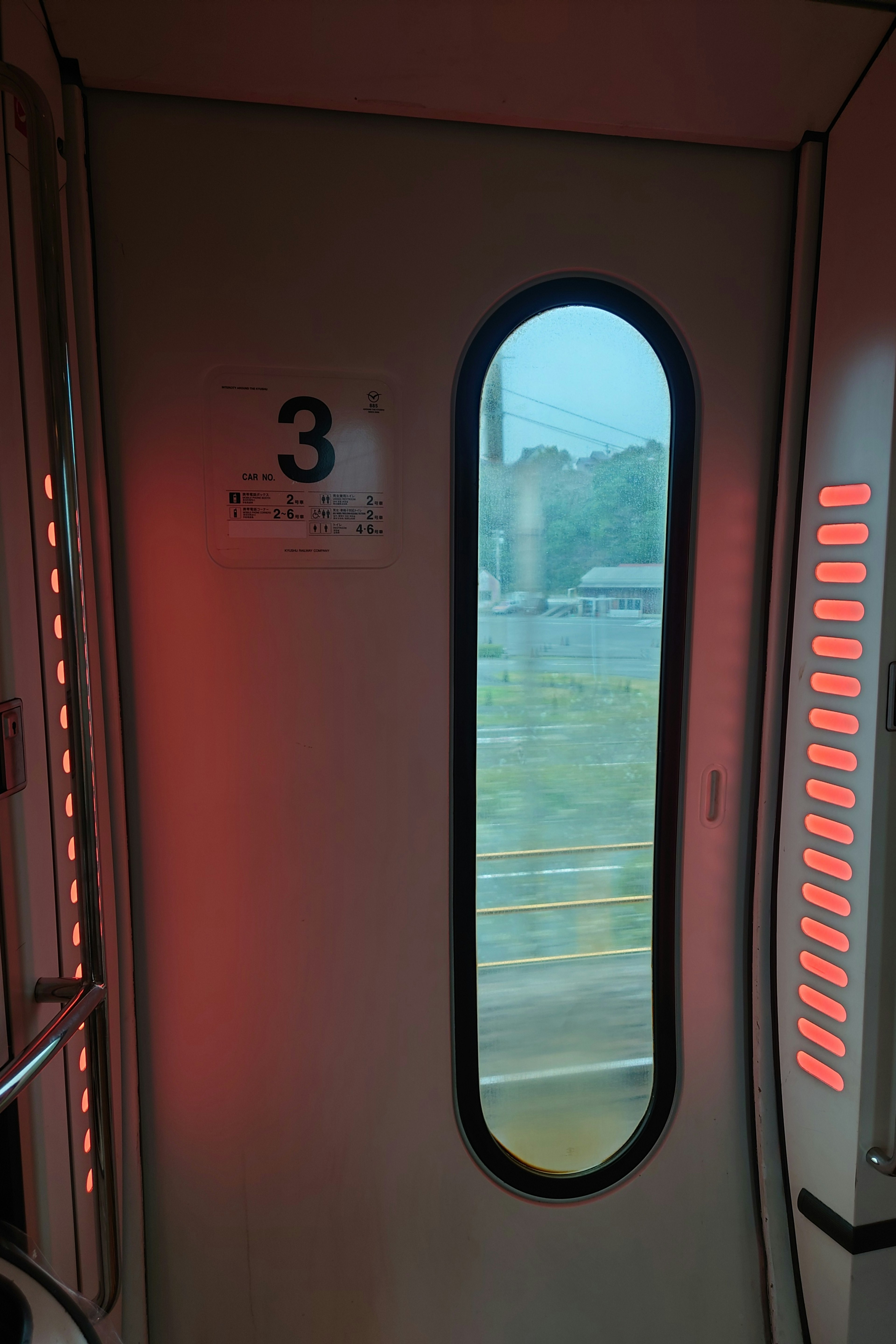 Vista interior de una puerta de tren con iluminación roja y una ventana redonda que muestra el paisaje exterior