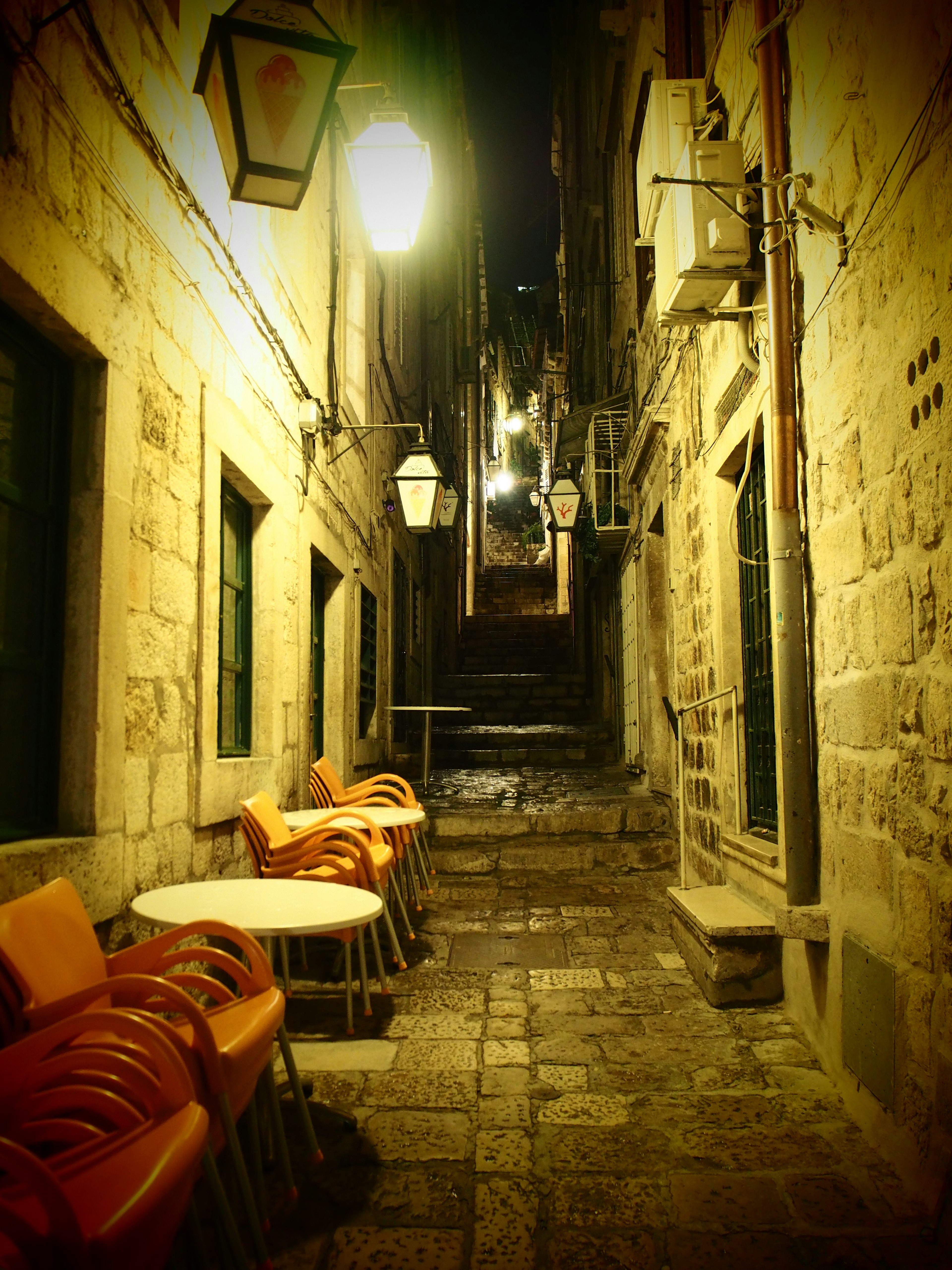 Enge Gasse bei Nacht mit gestapelten Stühlen und Tischen Steinmauern und Vintage-Straßenlaternen schaffen eine charmante Atmosphäre